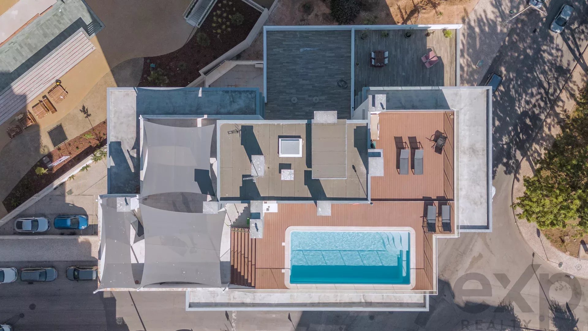 Aerial view of a modern rooftop with a rectangular turquoise pool, lounge chairs, and shaded seating areas on a wooden deck surrounded by walls and streets on three sides.