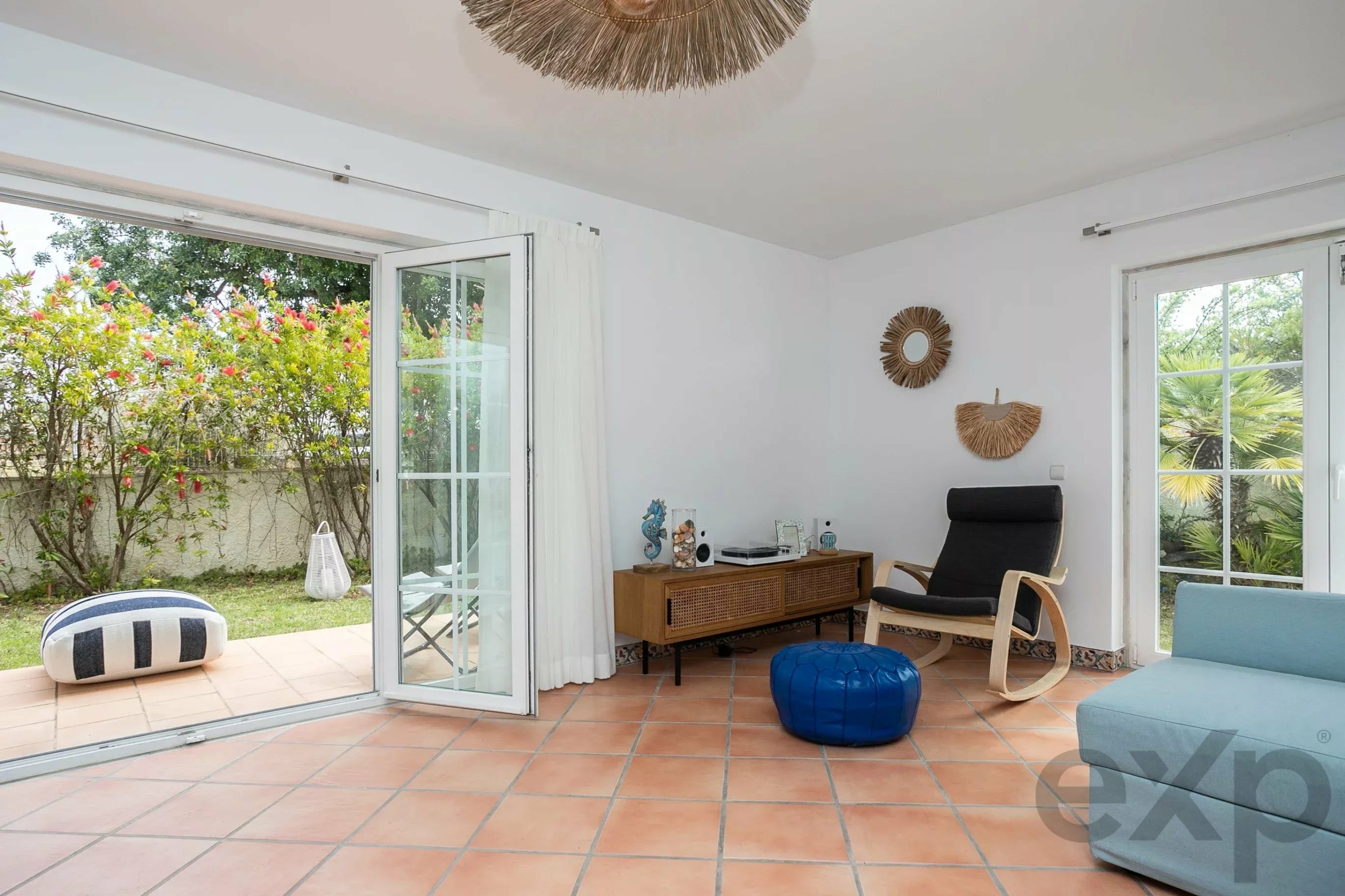 Bright living room with white walls and large glass doors opening to a sunny garden with pink flowers; wicker wall decor and a black lounge chair beside a wooden console.