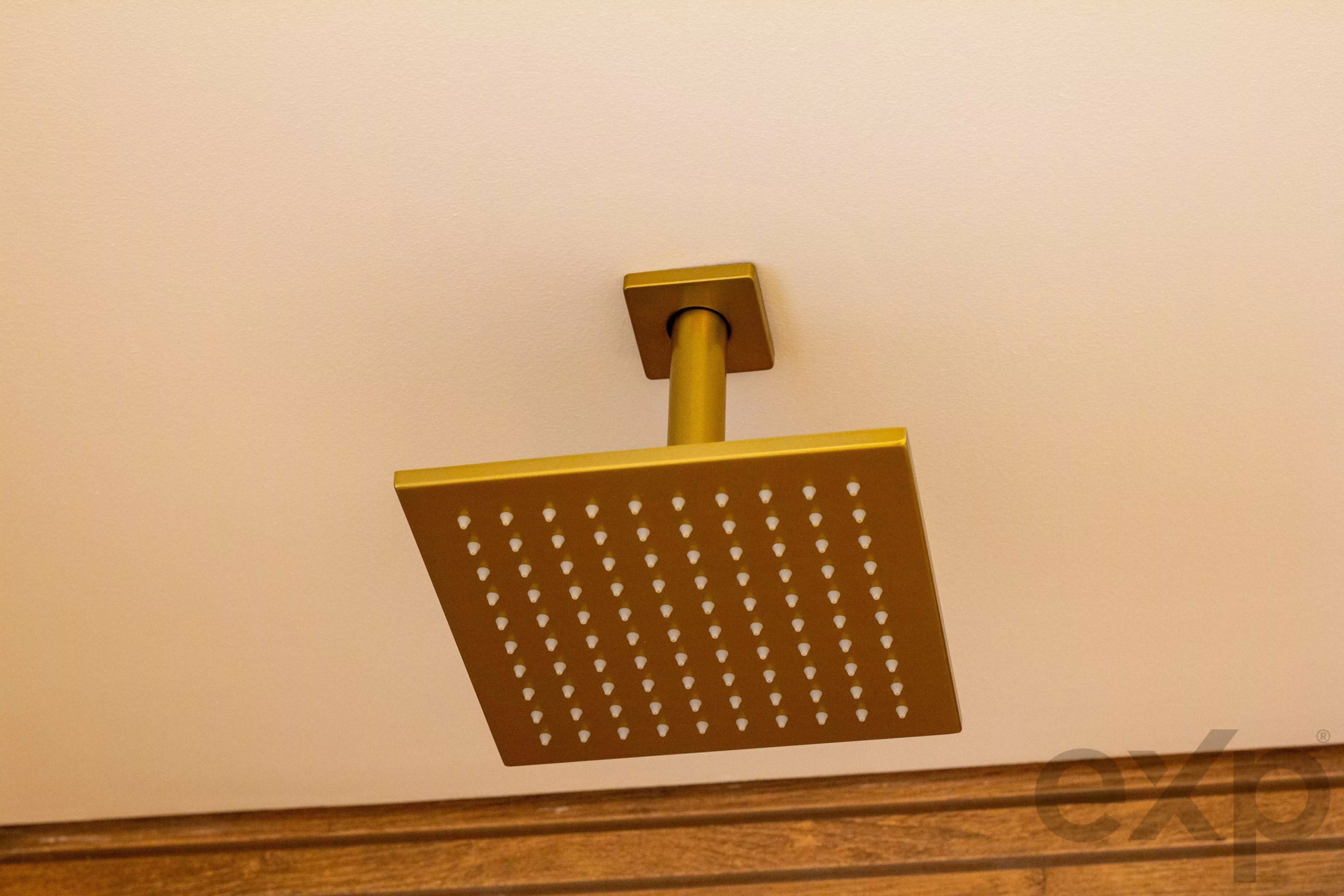Gold square rainfall shower head mounted on a beige wall, seen from below, with many water outlets visible.
