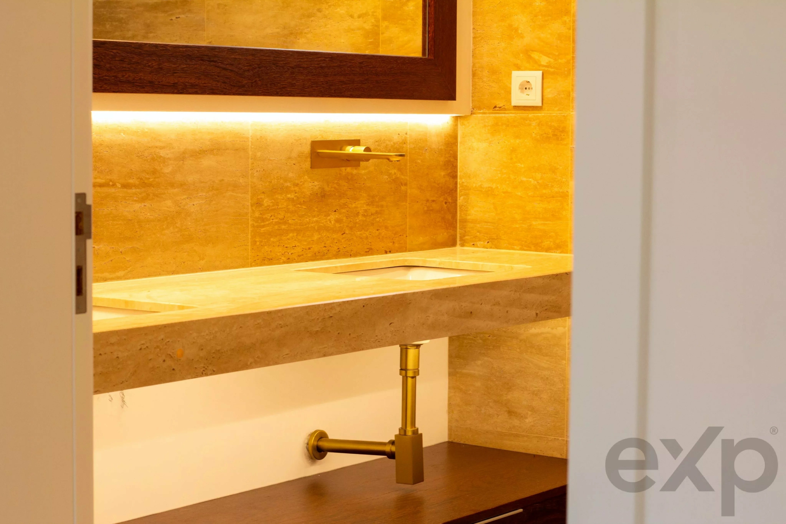 View of a bathroom sink with a gold faucet, beige marble countertops, and backlit tile backsplash seen through a doorway.