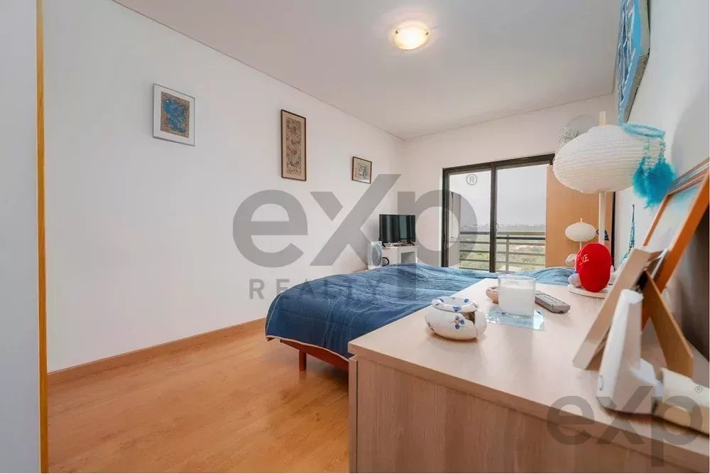 Bedroom with blue duvet, wooden floor, and dresser in the foreground; sliding balcony doors at the far end.