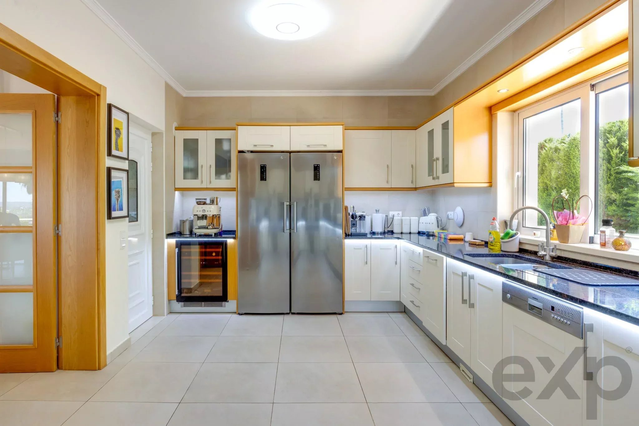 Bright modern kitchen with white cabinets, stainless steel double refrigerator, and a large window overlooking greenery at the right.
