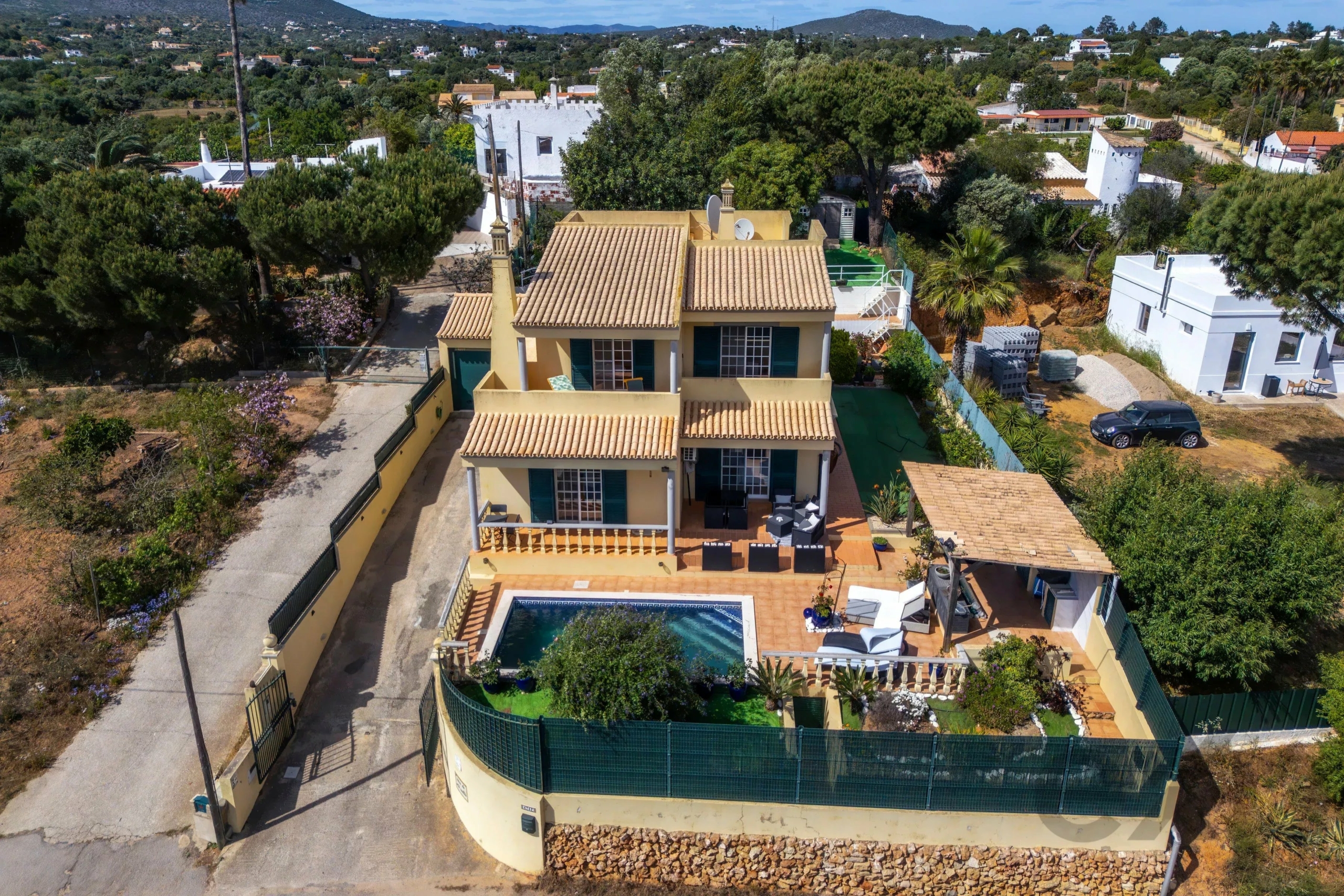 Aerial view of a cream-colored house with a terracotta tile roof, a backyard pool, and a fenced patio in a leafy hillside neighborhood (Spain/ Mediterranean style).