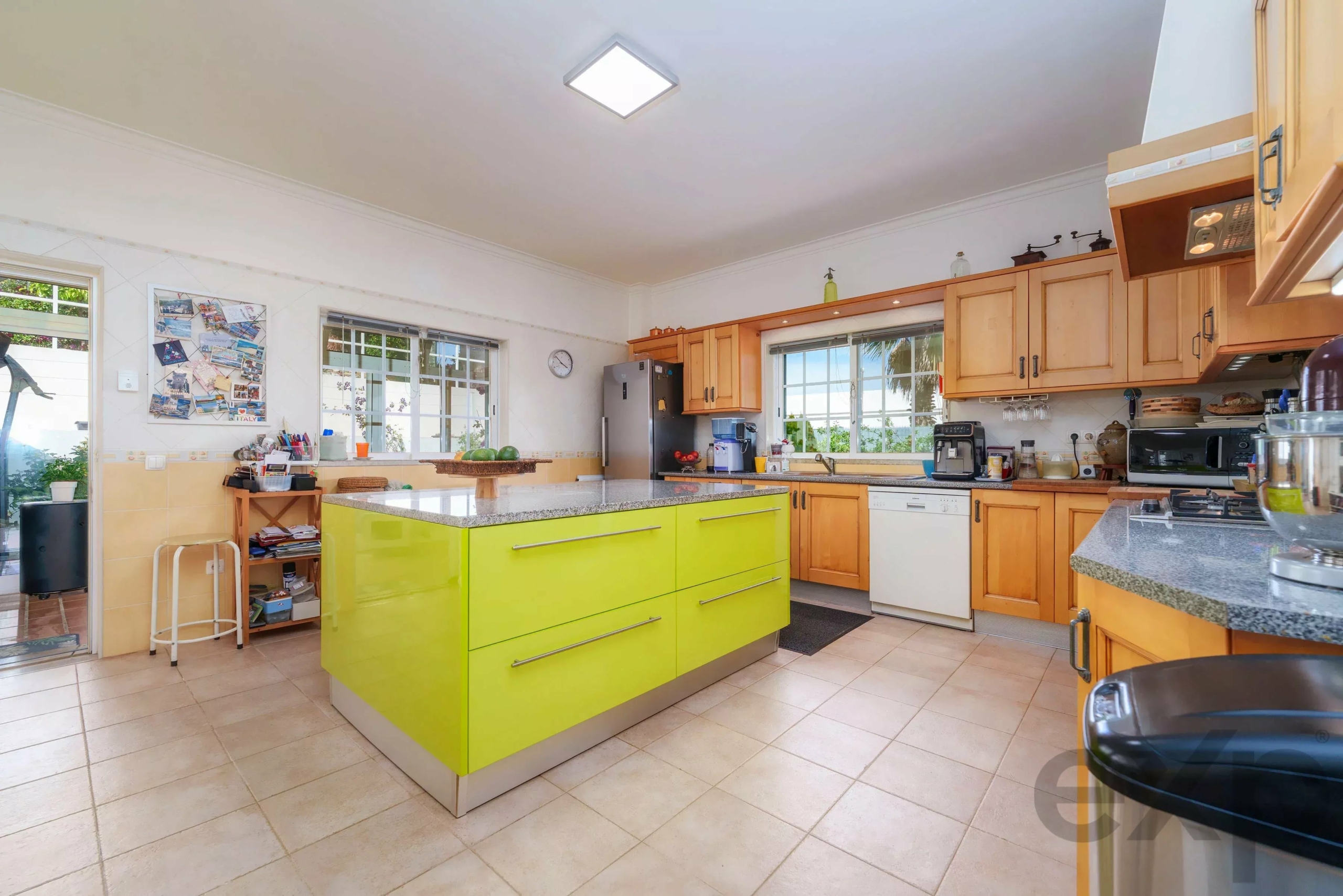 Bright kitchen with a lime-green central island, wooden cabinets, and stainless appliances under a high ceiling with large windows.