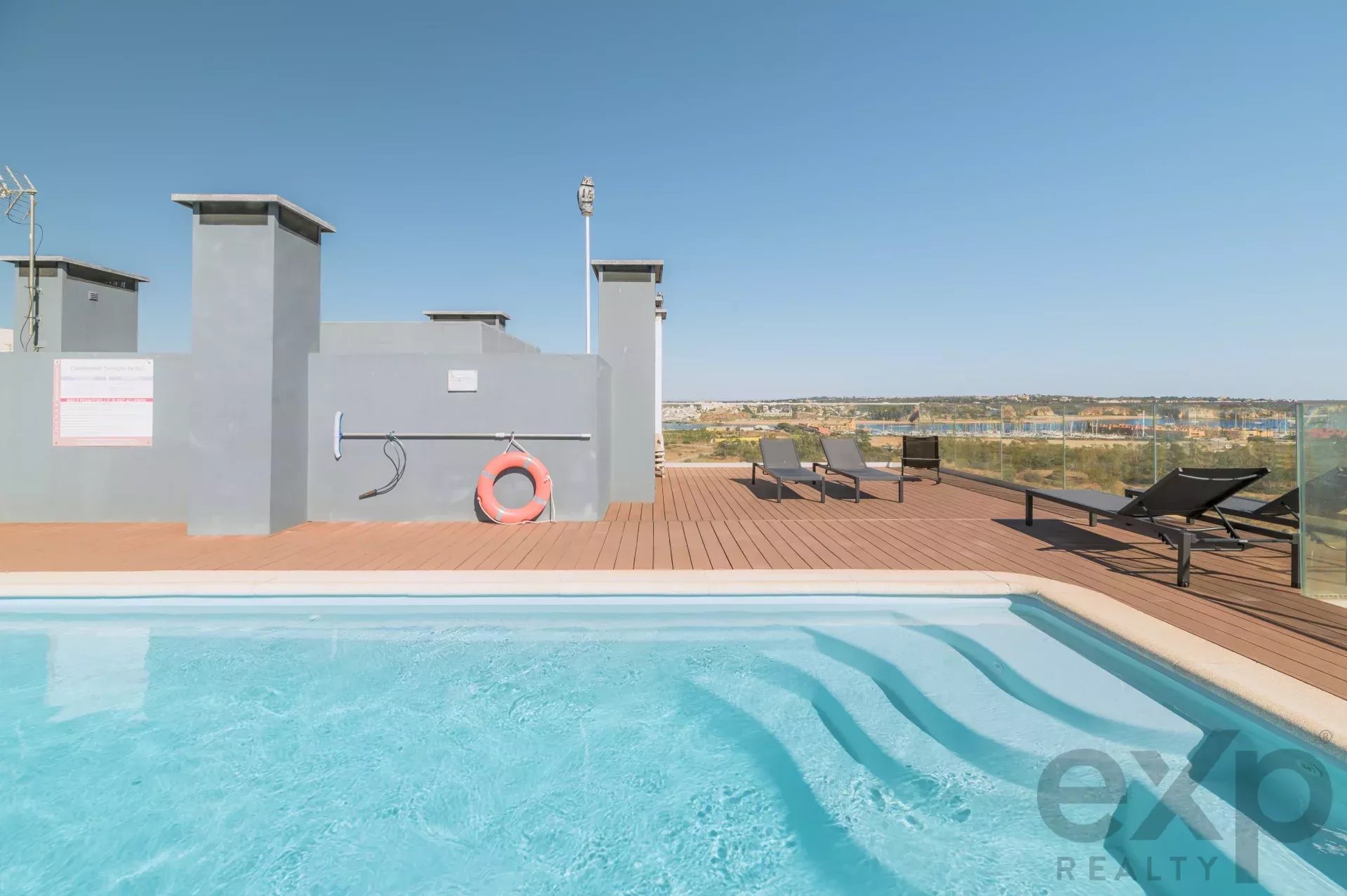 Rooftop pool with turquoise water, wooden deck, and several lounge chairs overlooking a distant landscape on a sunny day.