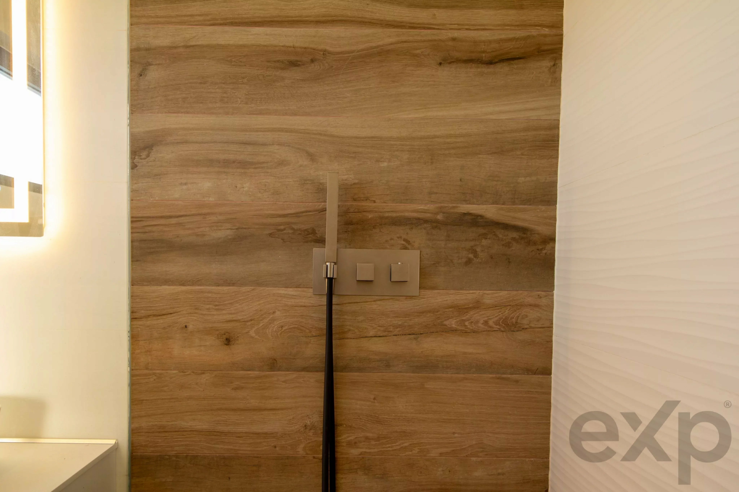 Wood-paneled wall with a beige outlet and two square switches mounted in the center; a vertical metal rod extends downward. Left edge shows a lit bathroom mirror, right edge a white textured wall.