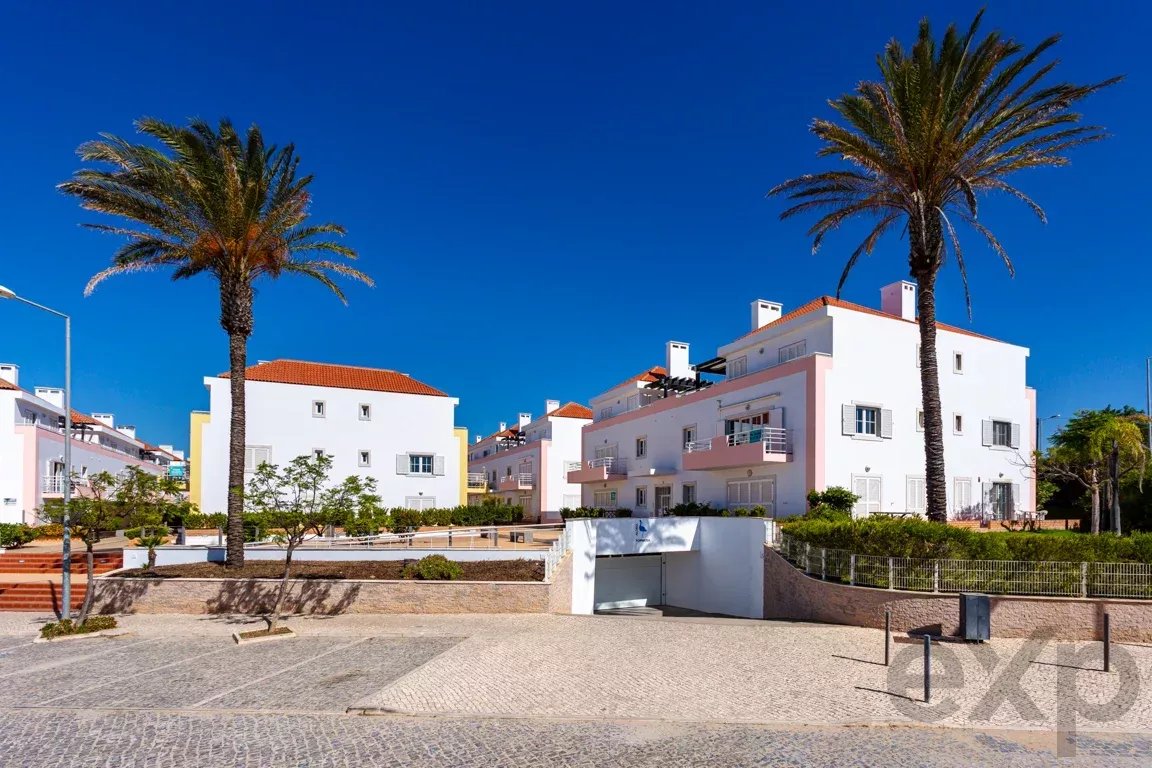 Sunny coastal plaza with white pastel apartment buildings, red-tiled roofs, and tall palm trees under a bright blue sky.