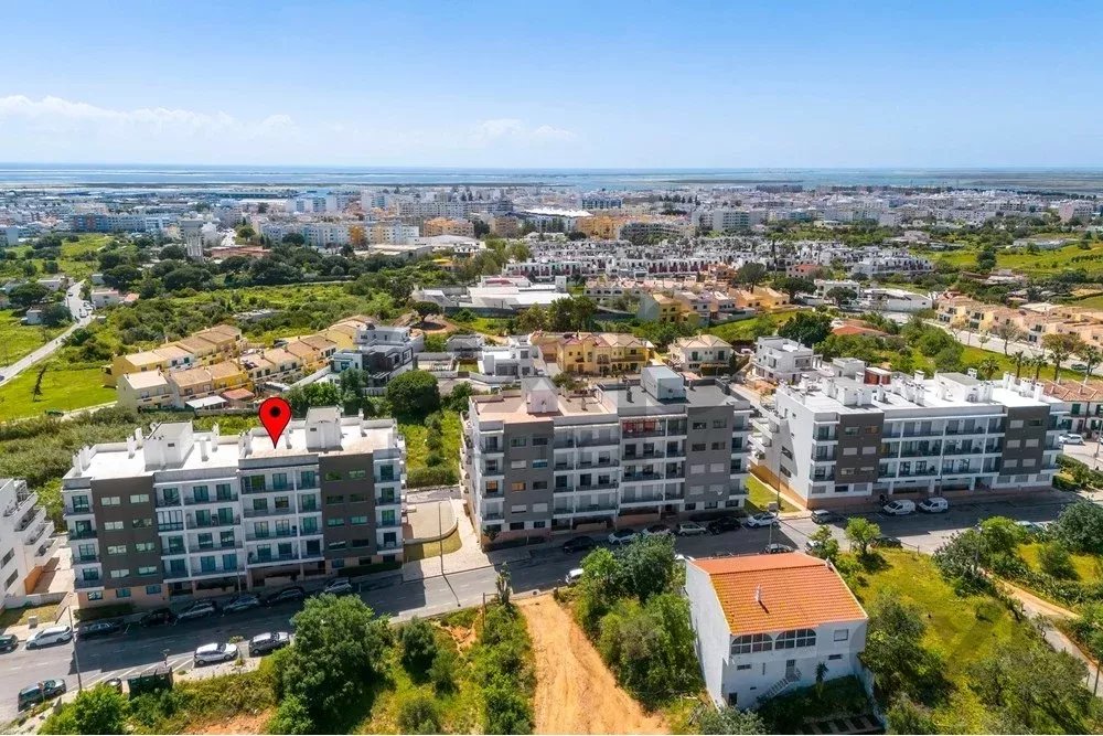 Aerial view of a residential complex with white and gray buildings; a red map pin marks one building, with a coastal town and sea in the distance.