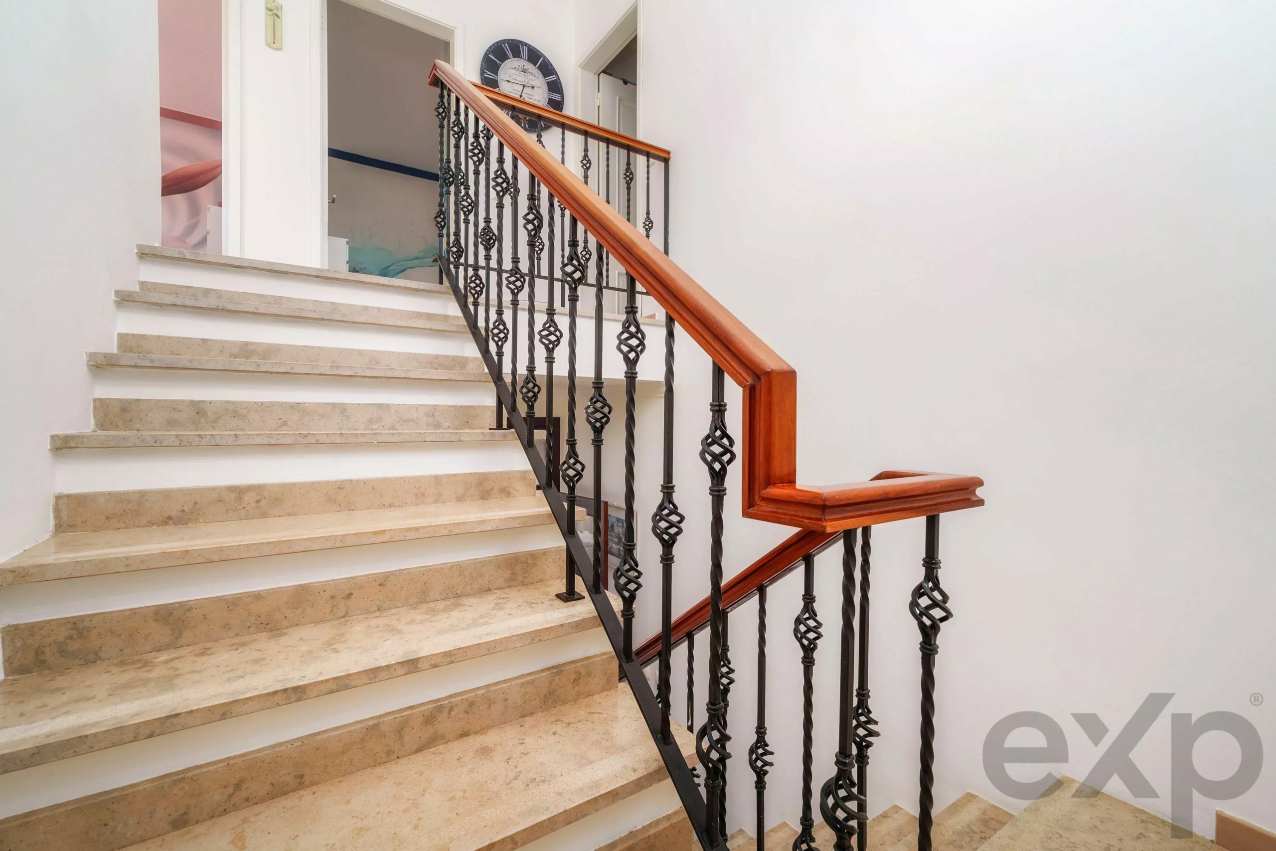 Interior stairwell with beige stone steps and decorative wrought-iron railing topped by a polished wooden handrail; a clock hangs on the landing wall above the stairs.