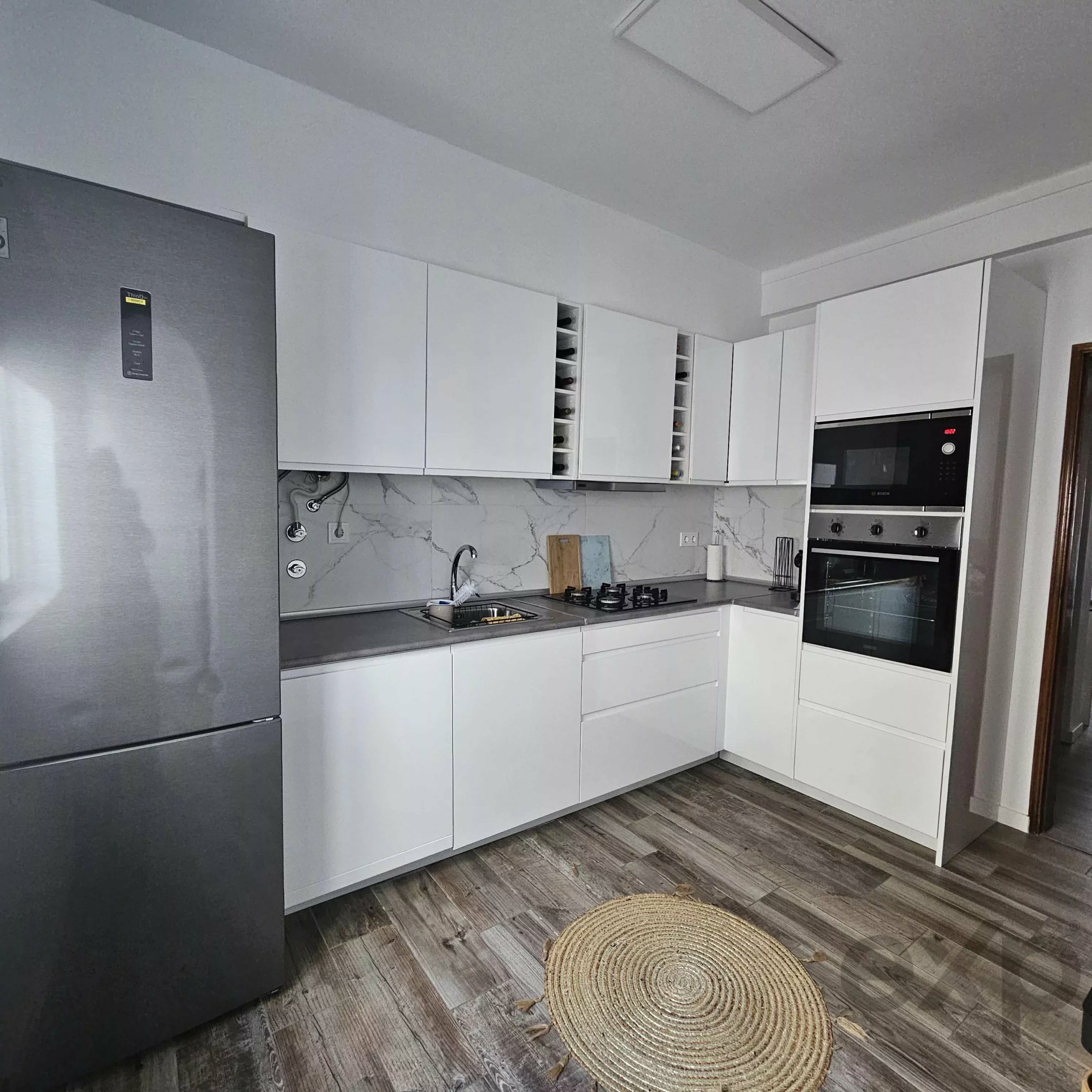 Modern kitchen with white cabinets and stainless steel fridge, built-in ovens, marble backsplash, and two round woven rugs on wood floor.