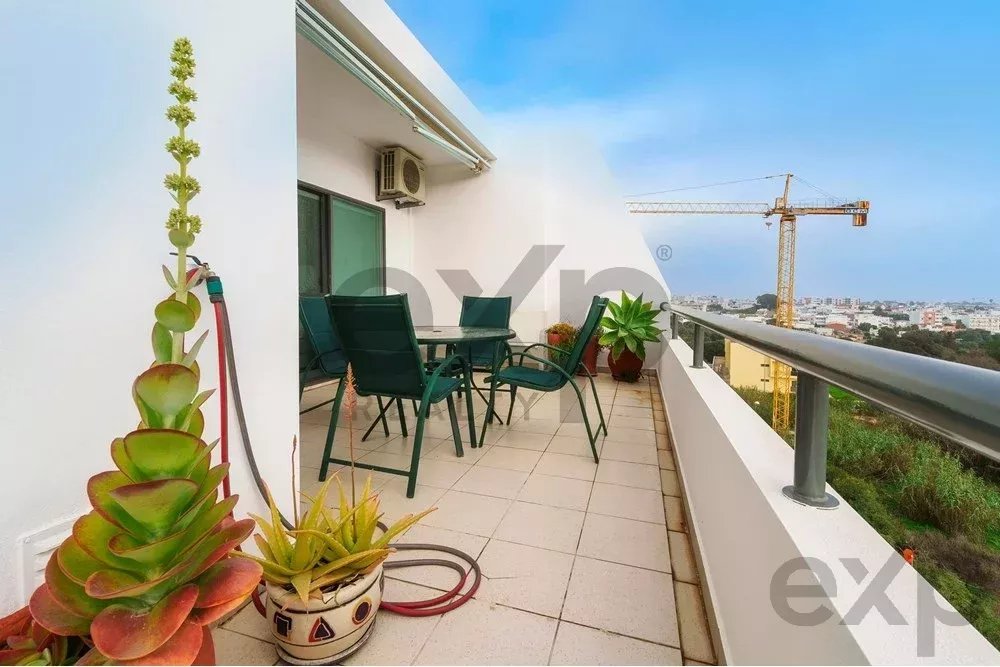 Balcony with a glass-table and green chairs, potted succulents, and a city skyline in the distance under a blue sky.