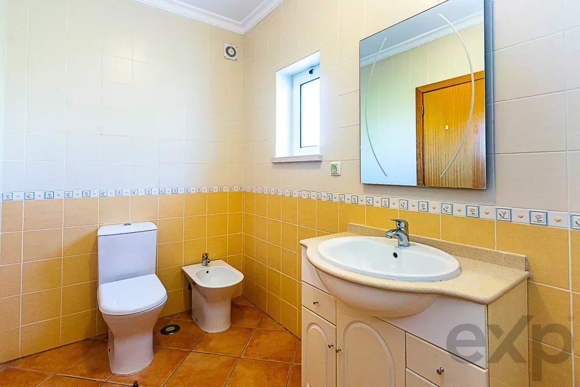 Bathroom with white toilet and bidet against yellow-tiled walls, beige countertop, and a large mirror above a curved sink cabinet.