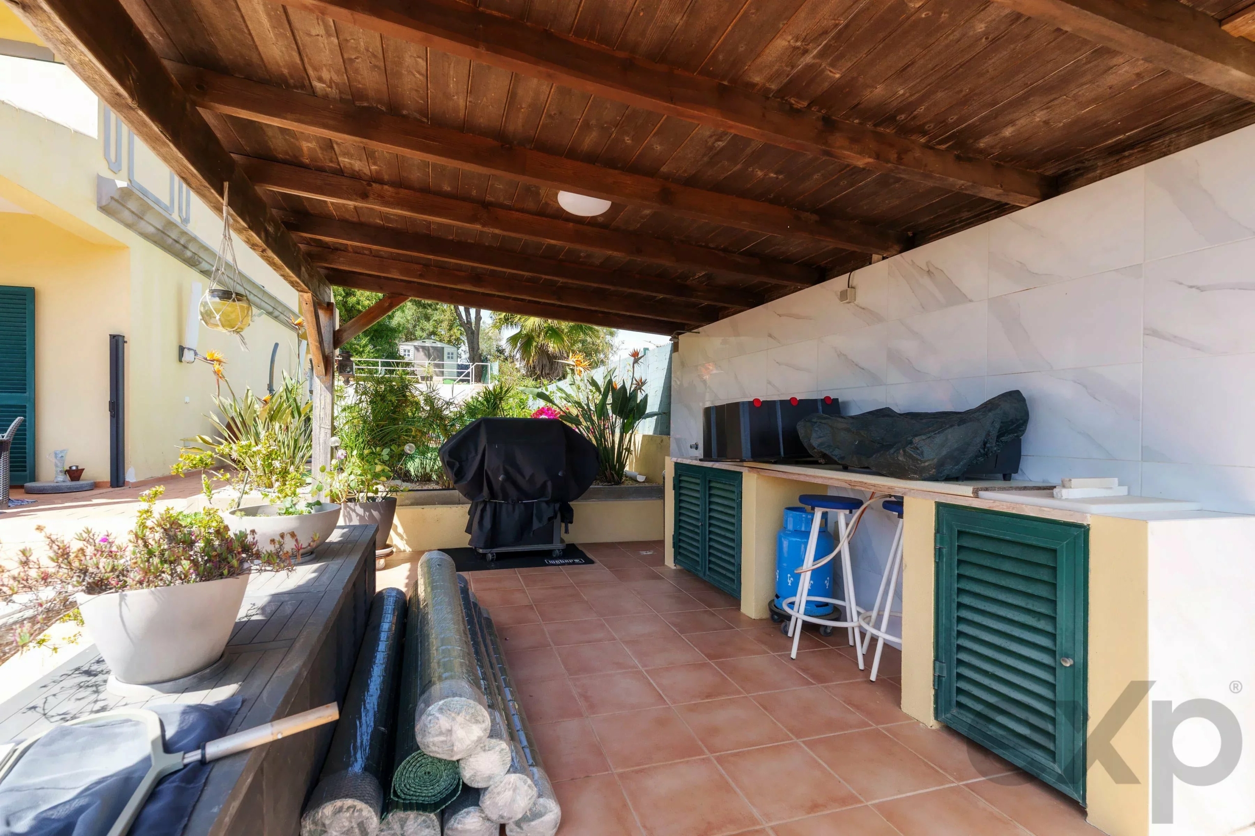Covered outdoor patio with a dark wooden beam ceiling, yellow walls, and potted plants along the edge near a grilling station with a tarp-covered grill.