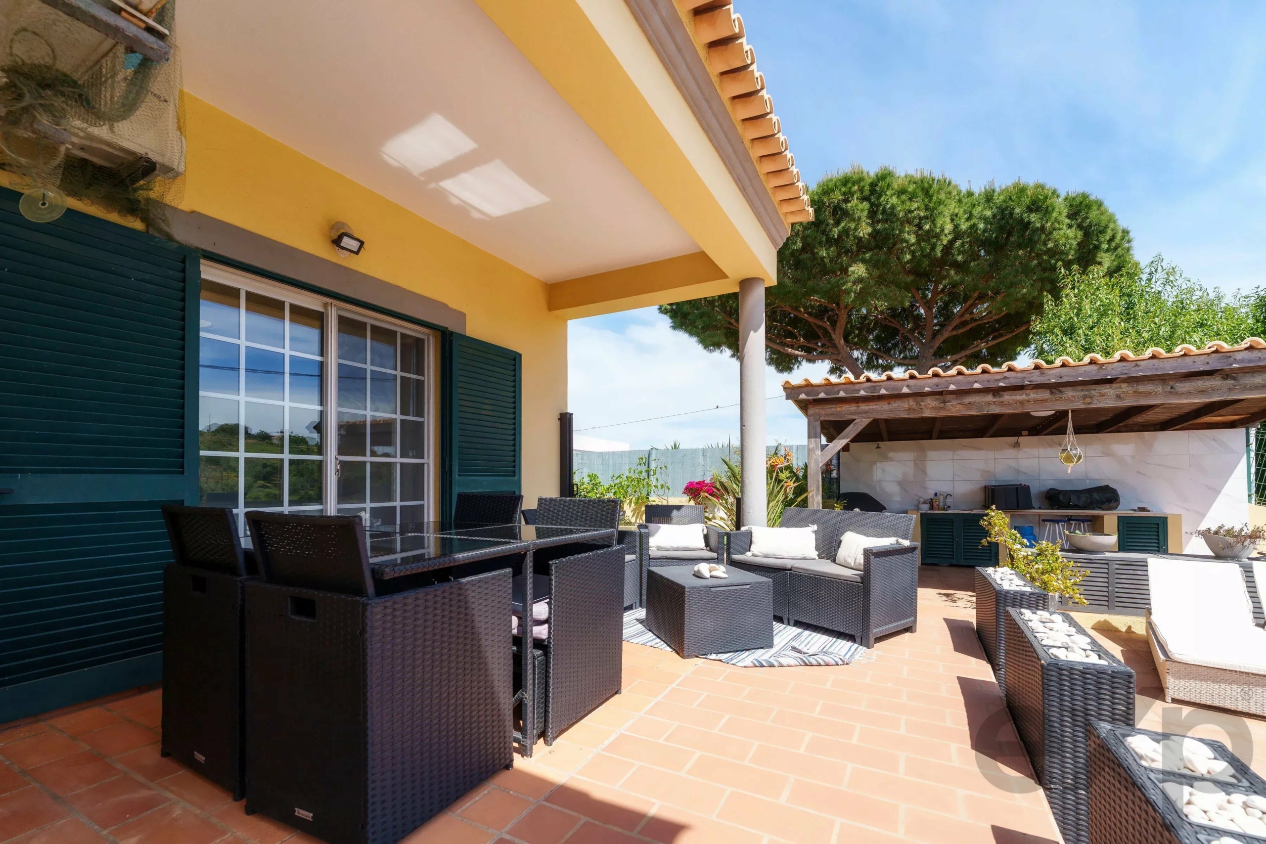 Sunny outdoor patio with dark wicker seating, yellow walls, and a large green tree in the background.