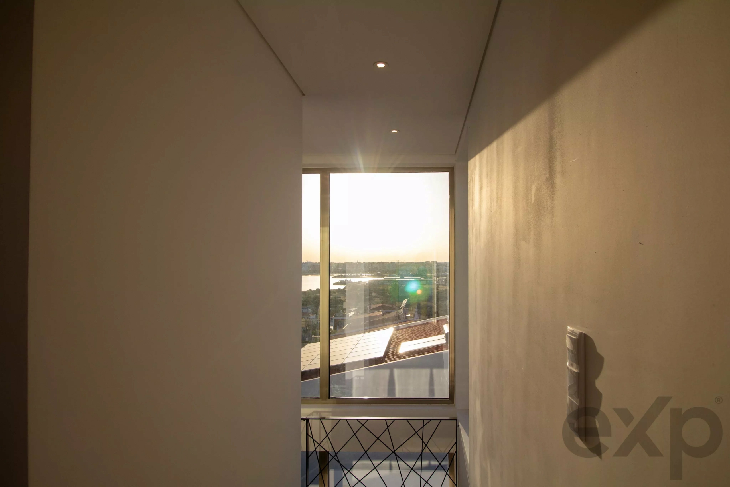 Narrow hallway heading toward a large window with sunlight and a balcony outside.
