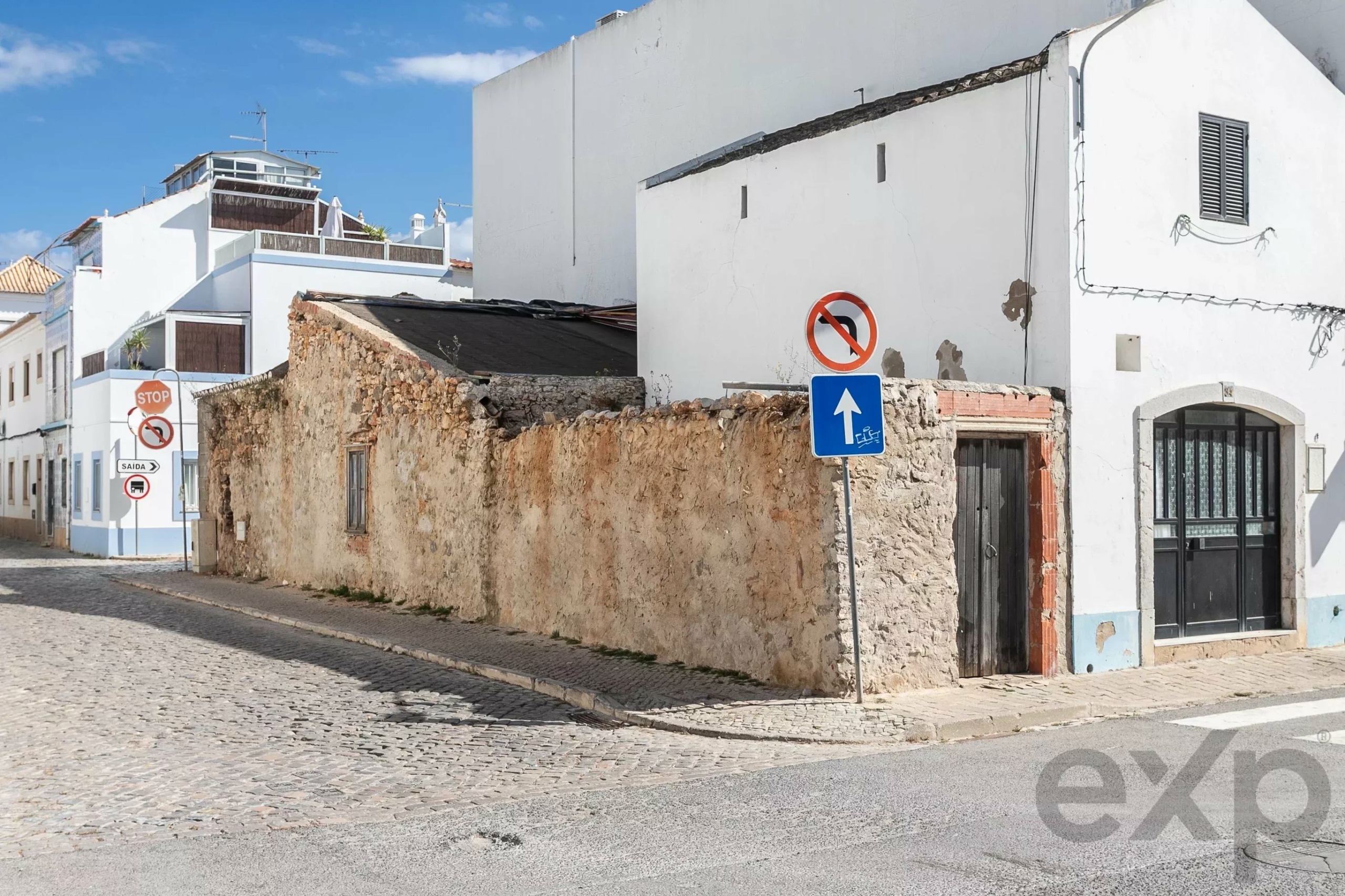 Cobbled street beside white buildings with a rough stone wall and traffic signs in the foreground (no entry and blue upright-arrow signs).