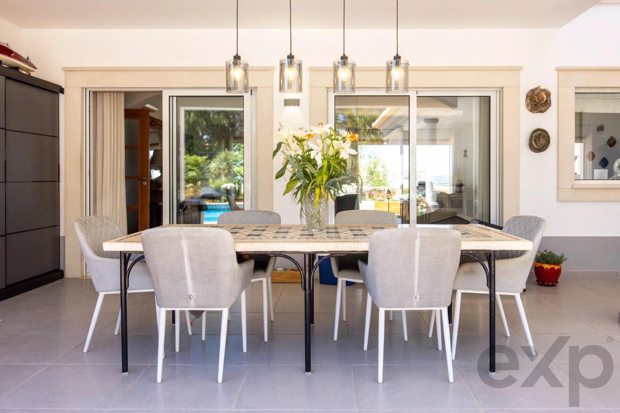 Outdoor dining area with a mosaic table and eight gray chairs, a vase of lilies, and glass sliding doors to a sunny backyard patio with hanging pendant lights.