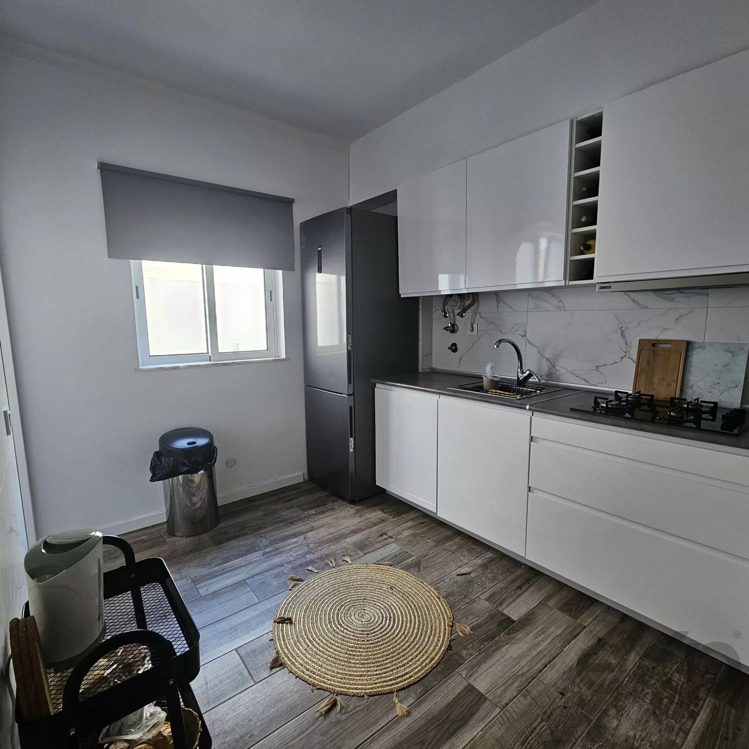 Modern kitchen with white glossy cabinets, marble backsplash, and stainless steel appliances, plus a circular braided rug on gray wood-look flooring.