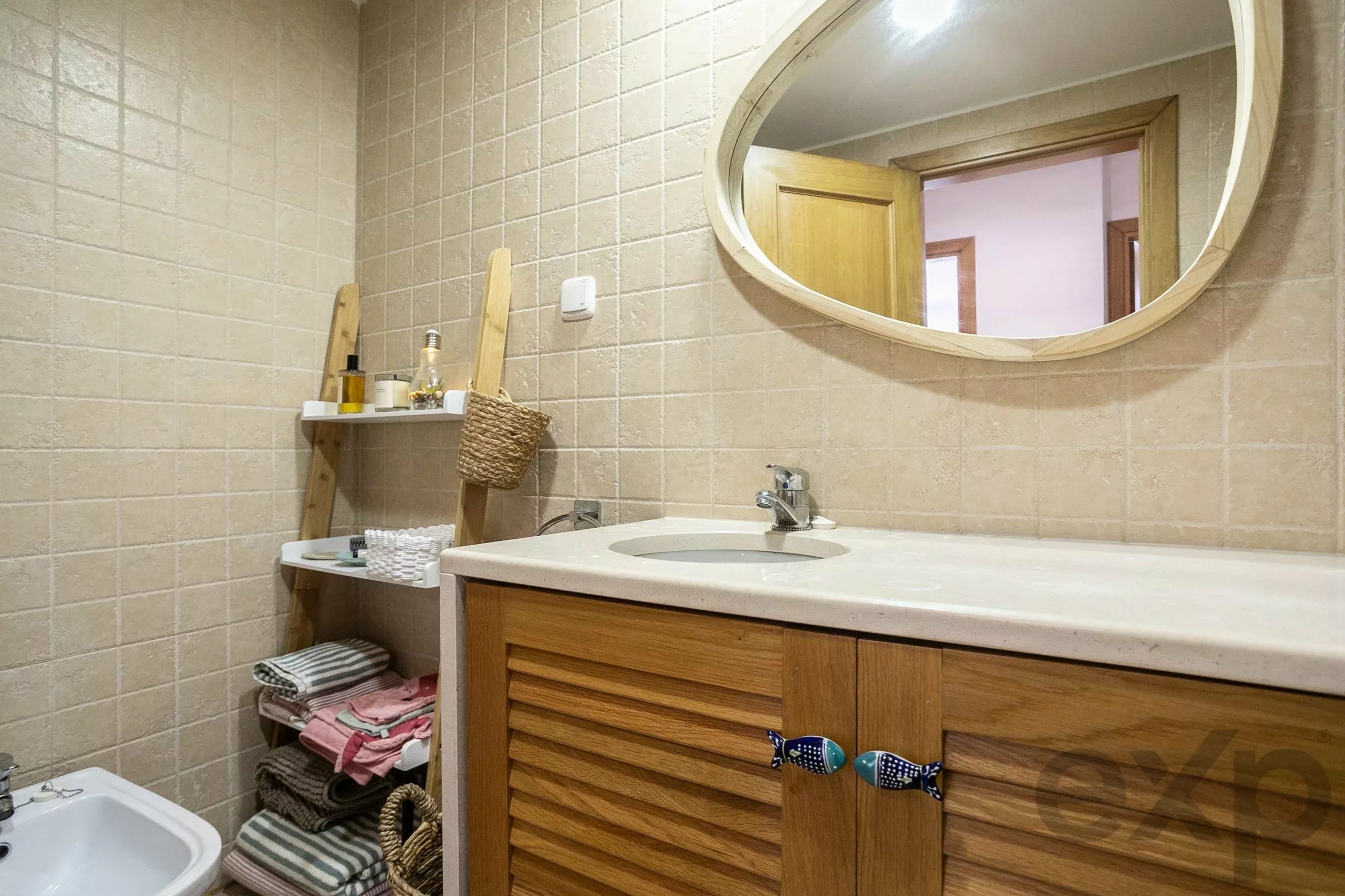 Beige-tiled bathroom with a round wooden-framed mirror above a marble countertop sink and stacked towels on shelves nearby.