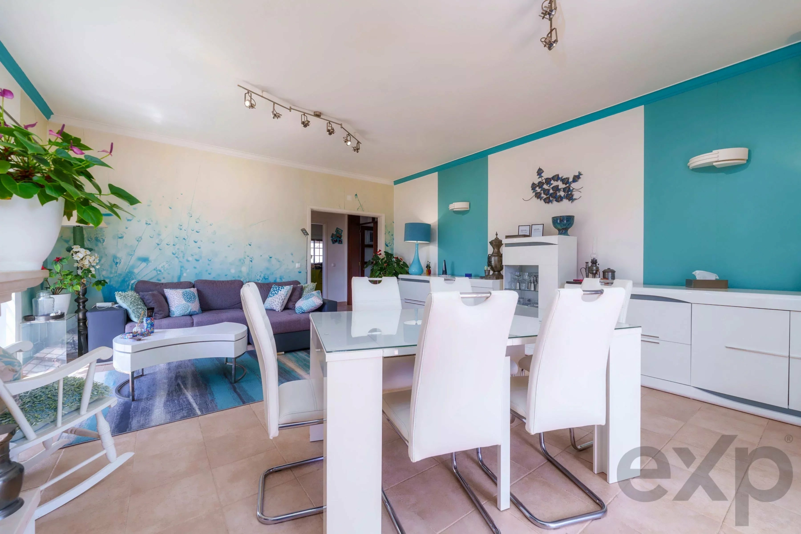 Open-plan living and dining room with white furniture, teal accent walls, and a glass dining table surrounded by white chairs.
