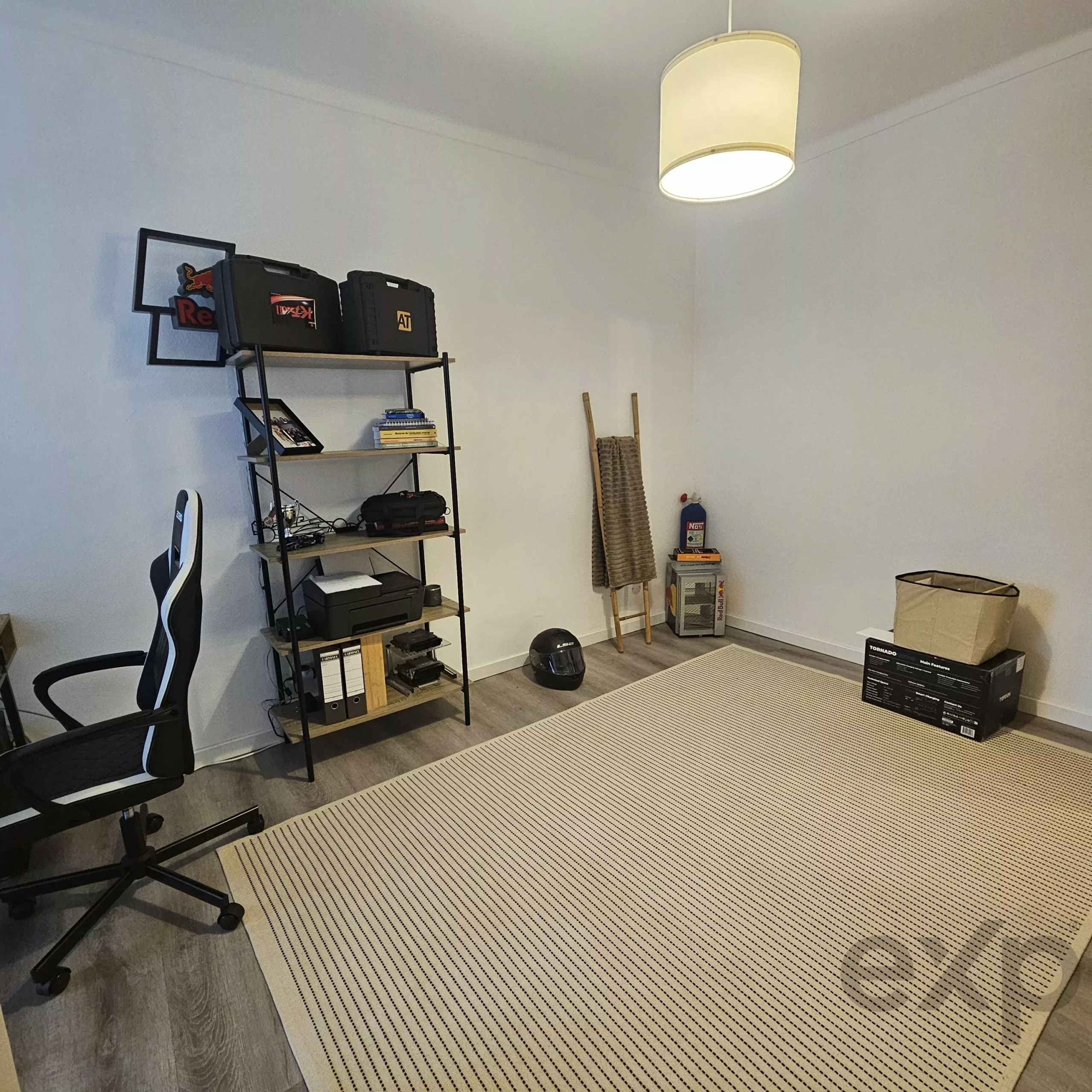 Corner workspace with a tall black shelving unit holding tool cases, boxes, and electronics; beige striped rug on a wooden floor and a large ceiling drum light overhead.