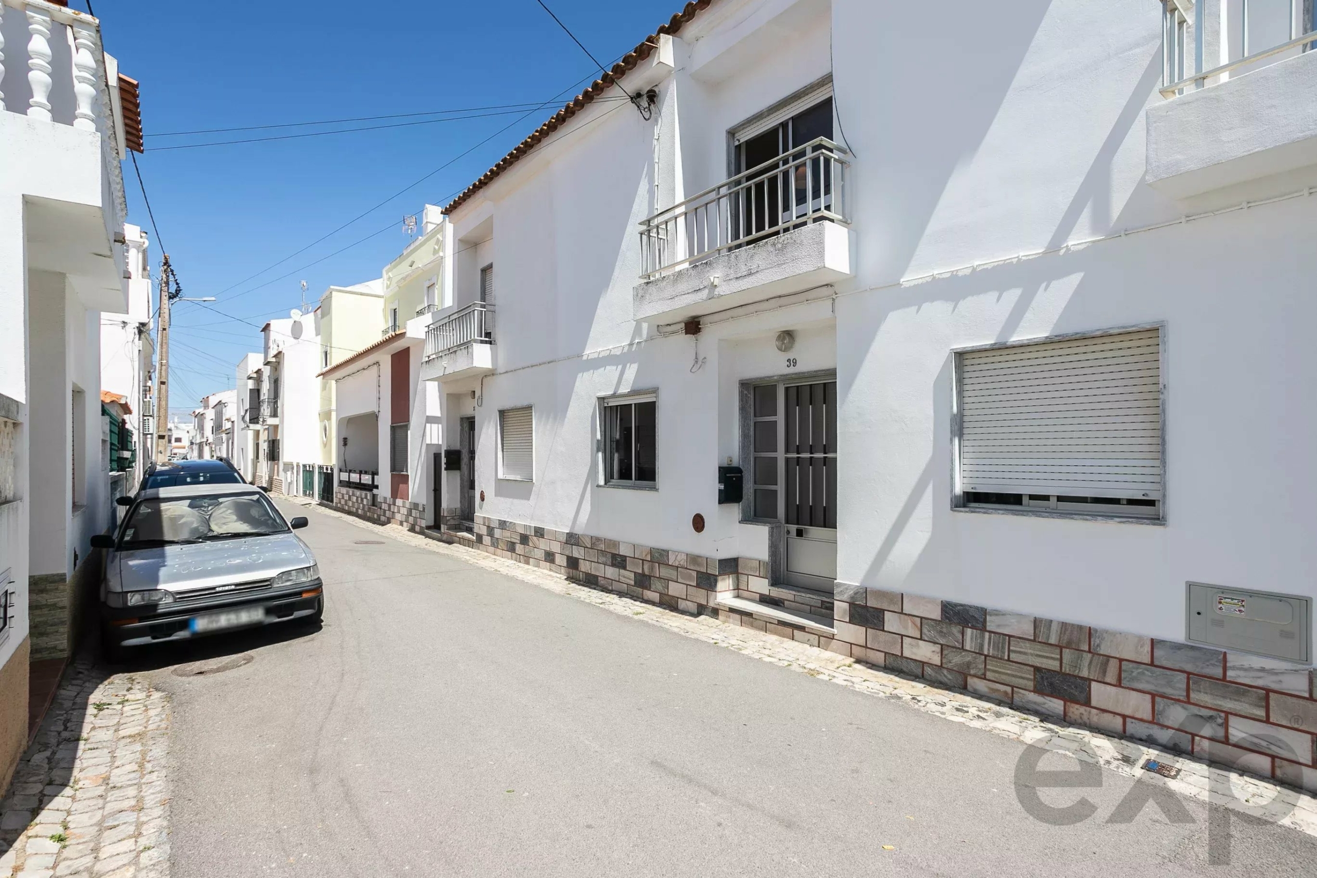 Narrow sunlit street lined with white houses, balconies, and shuttered windows; a silver car parked on the left.