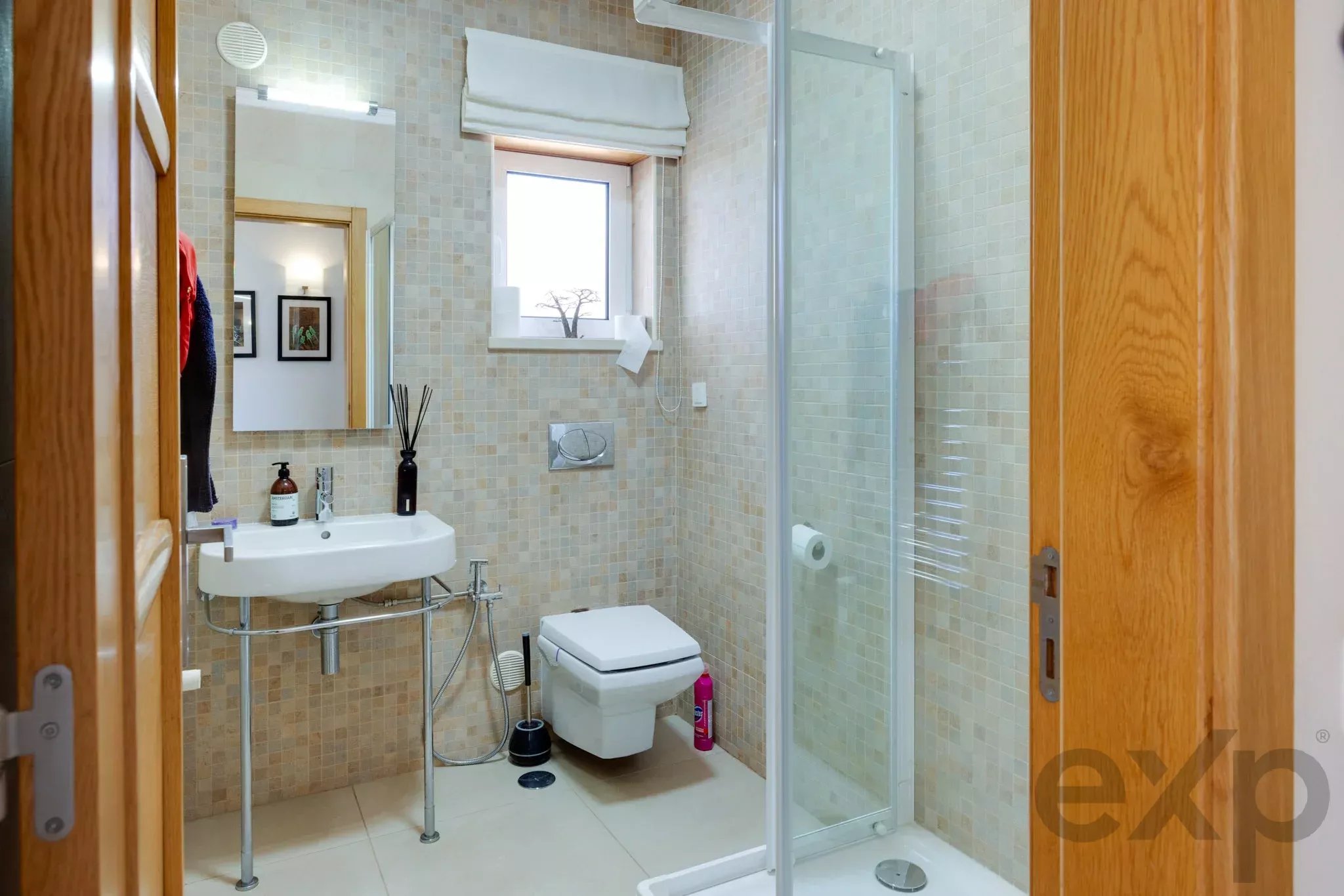 Small tiled bathroom with a white sink, wall mirror, toilet, and glass shower enclosure; wooden door frame visible.