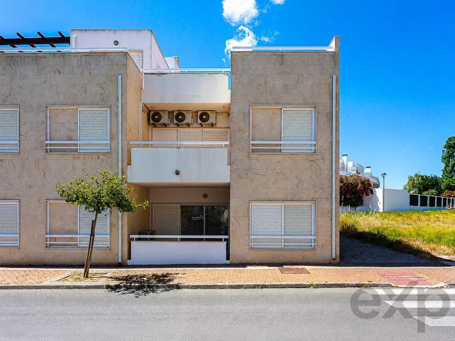 Two-story beige apartment building with balconies and shuttered windows under a bright blue sky.