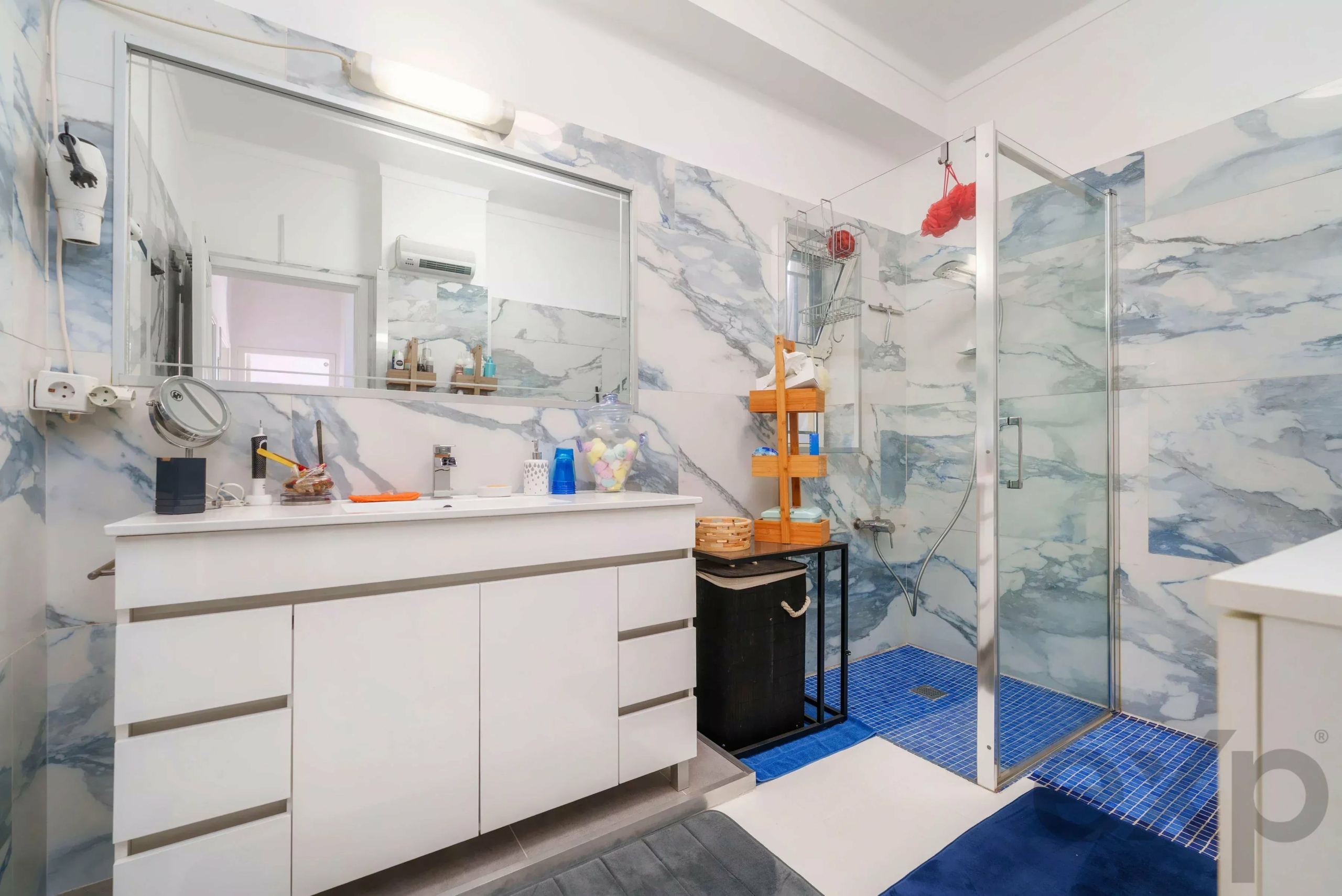 Modern bathroom with white vanity, marble-patterned walls, and a glass shower enclosure with blue tiled floor.