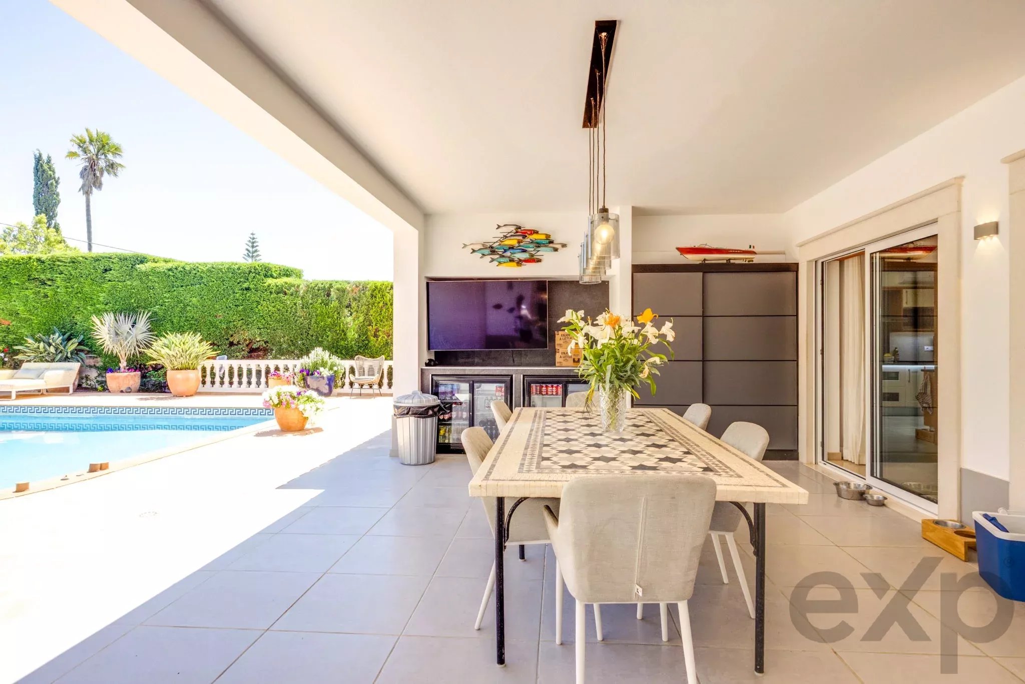 Outdoor covered patio with a long dining table and beige chairs, pool and green hedge in the yard beyond