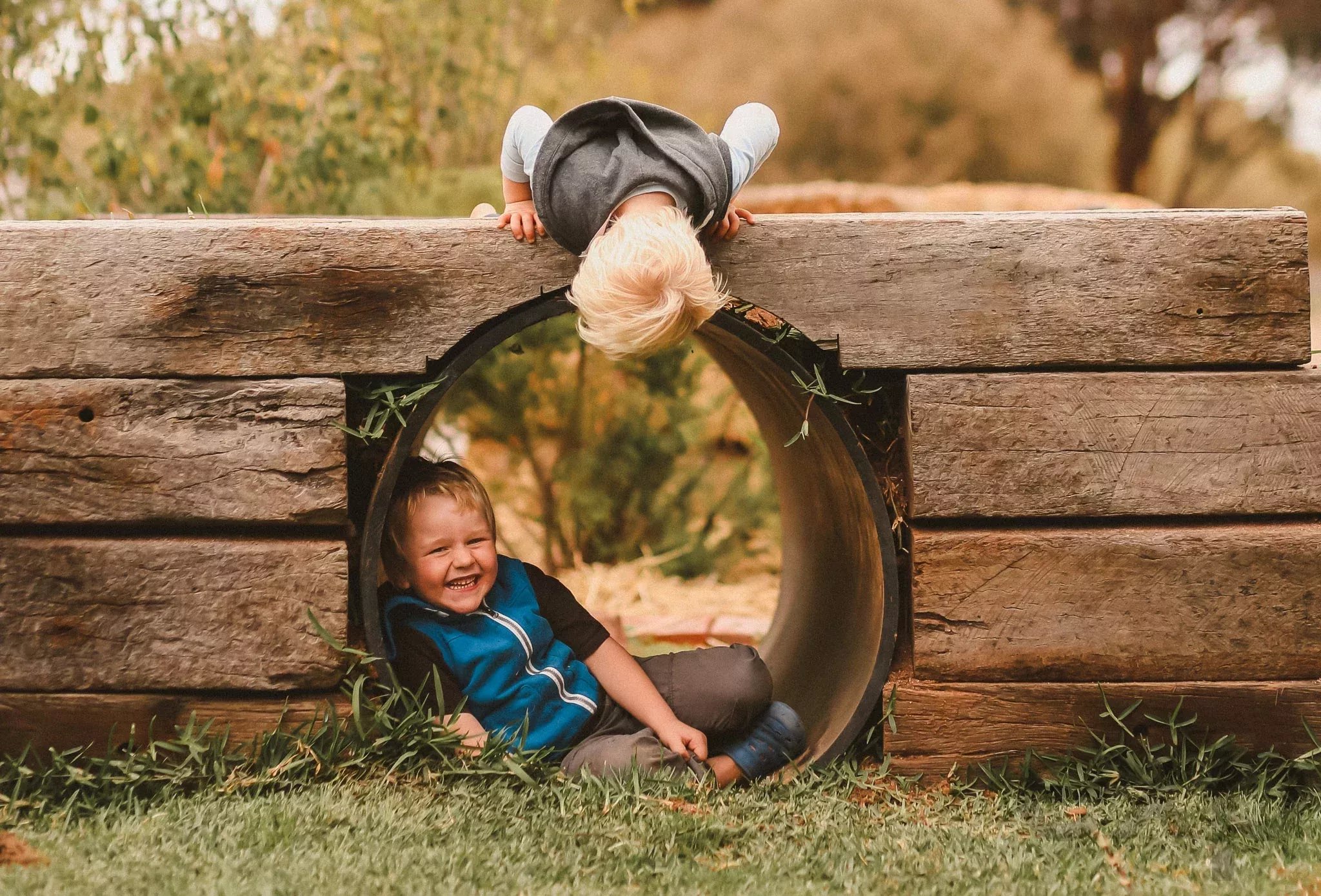 Two children playing in a wooden tunnel playground outdoors; one sits smiling inside while another leans over the opening above him.