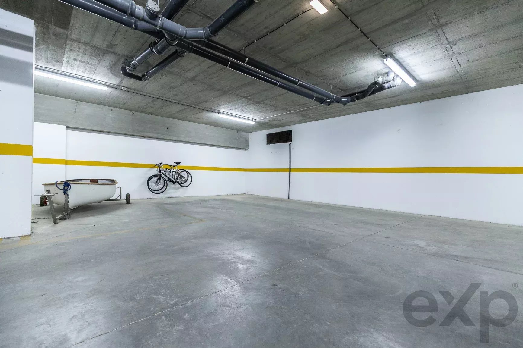 Underground parking area with a white wall and a yellow stripe, two bicycles hung on the wall, and a small boat on a trailer nearby.