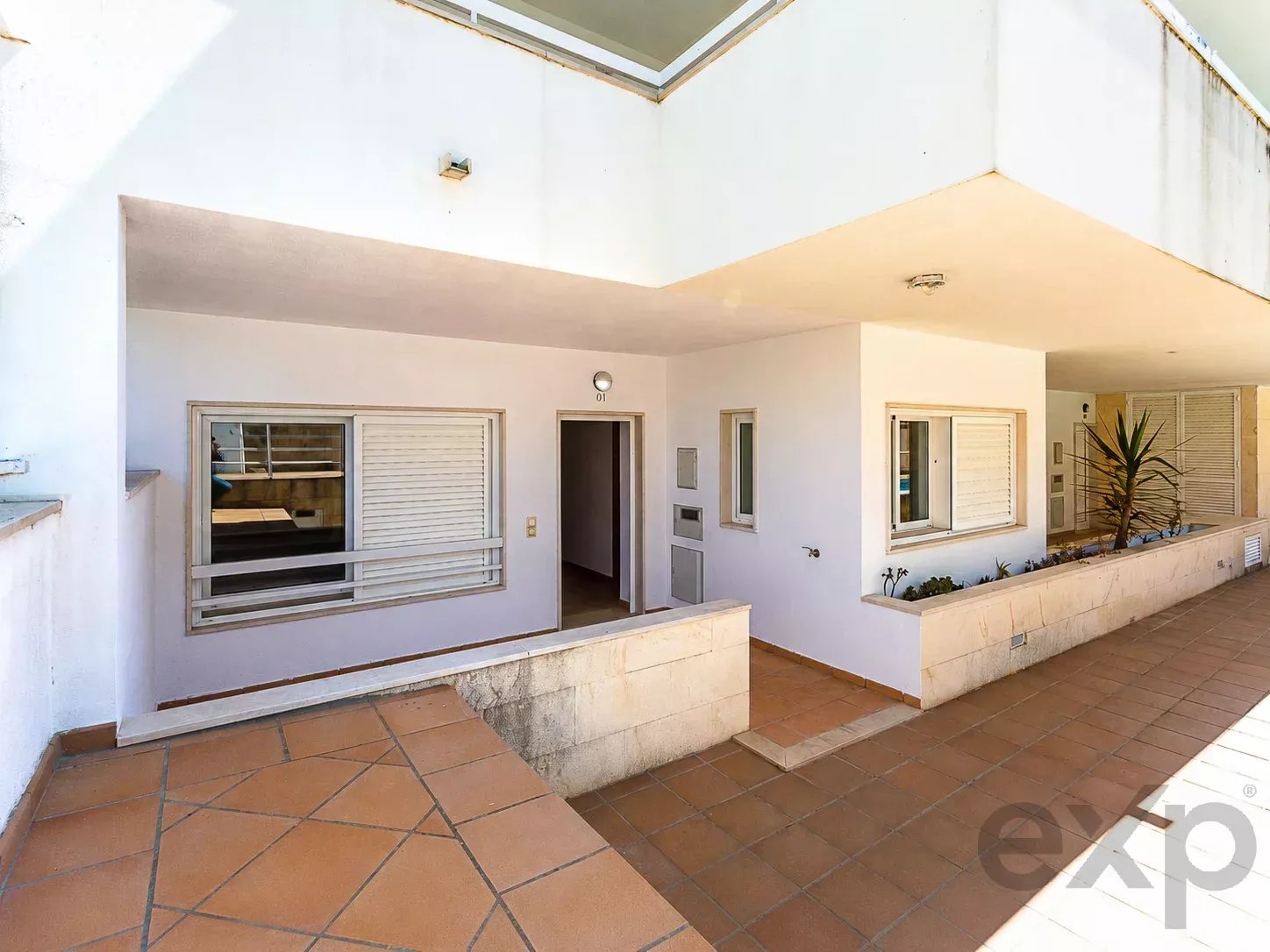 White modern building exterior with a covered walkway and tan tiled patio. A window with shutters is visible beside an open doorway (unit 01).
