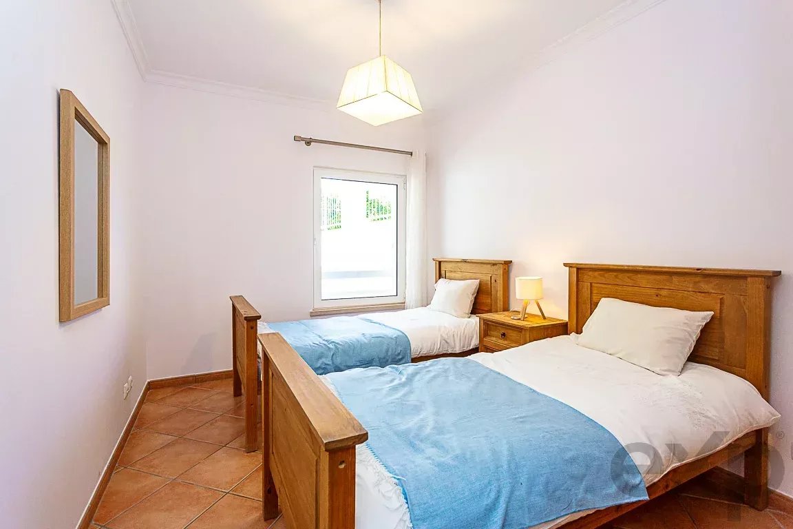 Twin bedroom with two single wooden beds, blue bed runners, white pillows, and a small wooden bedside table with a lamp between them; window at the far wall.