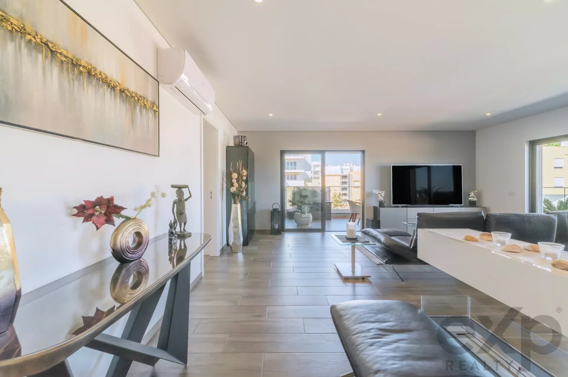 Modern living room with a gray sectional sofa, glass coffee table, wall art, and a large flat-screen TV near sliding balcony doors.