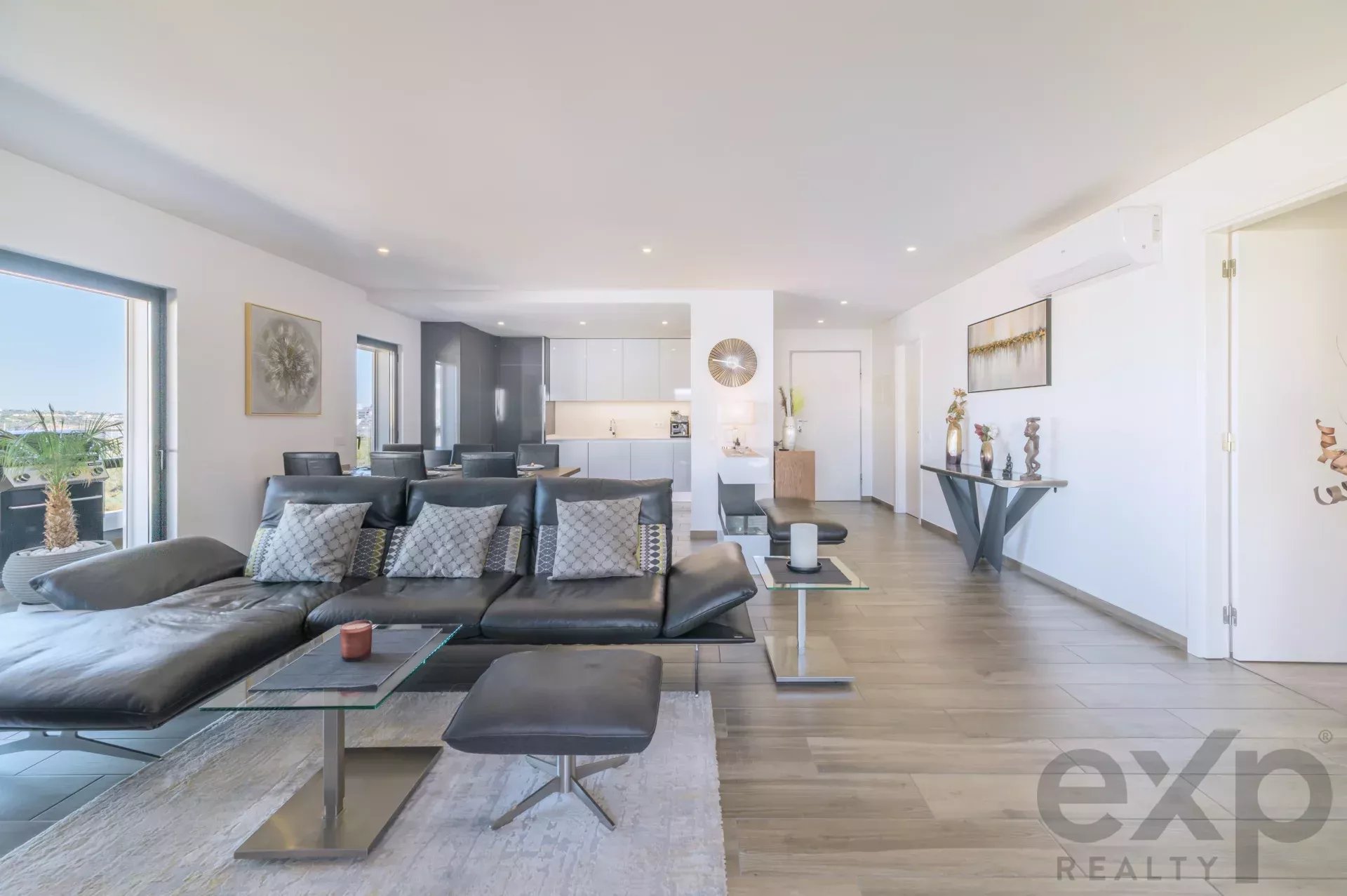 Open-concept living room with black leather sofa, glass coffee tables, neutral decor and a bright kitchen in the background.