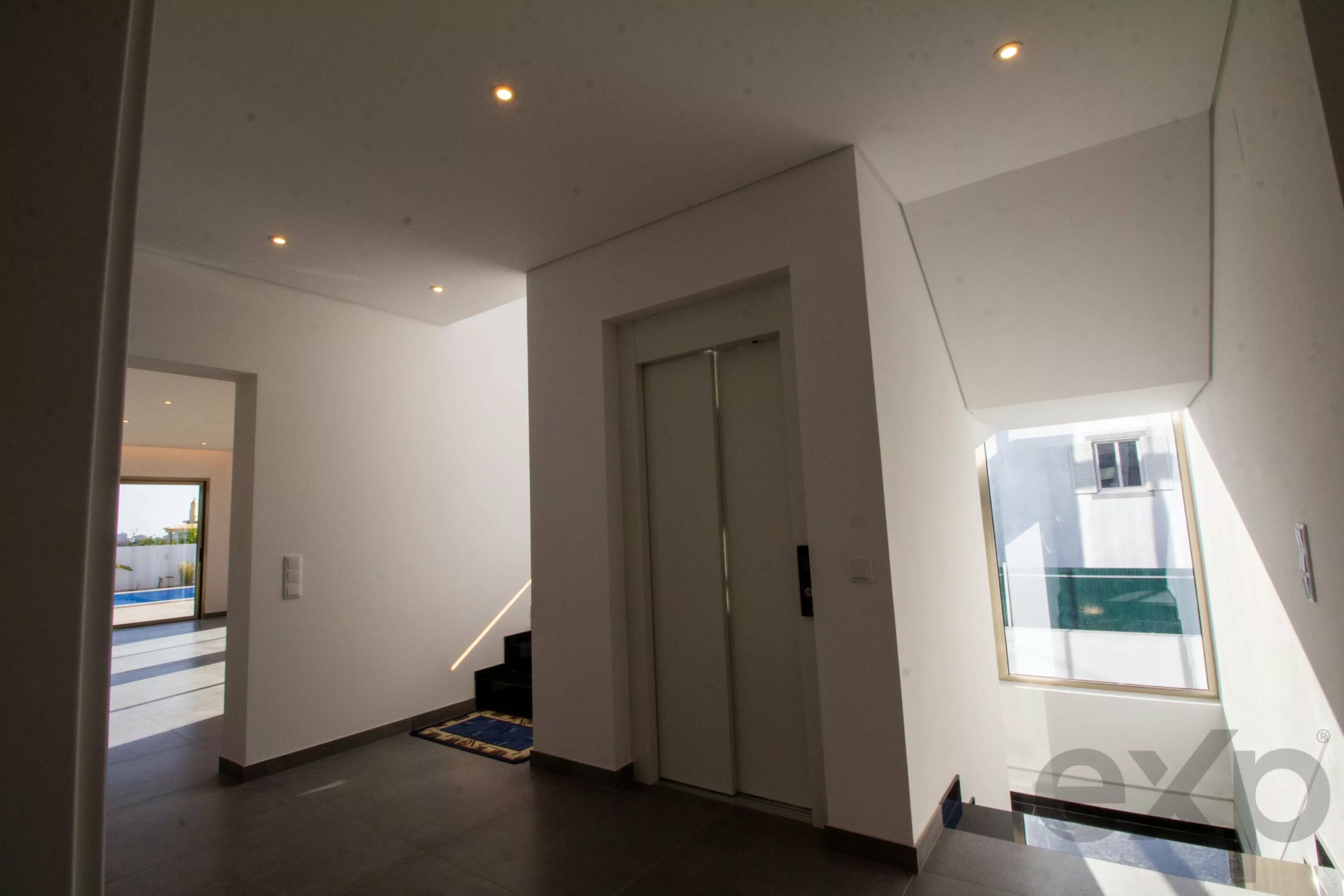 Modern townhouse foyer with a central elevator, a left-side staircase, white walls, and natural daylight from large windows.
