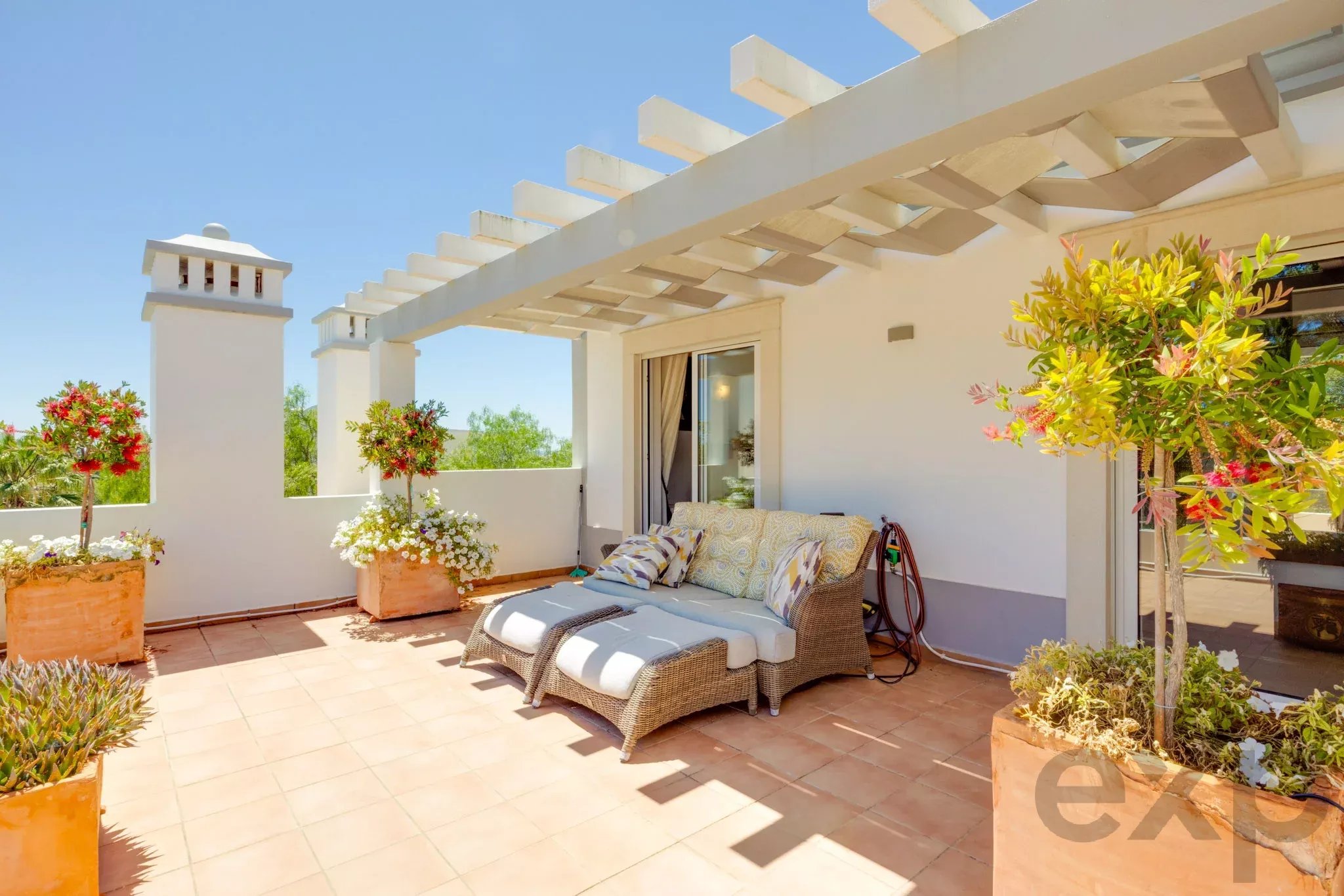 Sunny outdoor terrace with white pergola, beige tiled floor, wicker lounge chairs and potted plants.