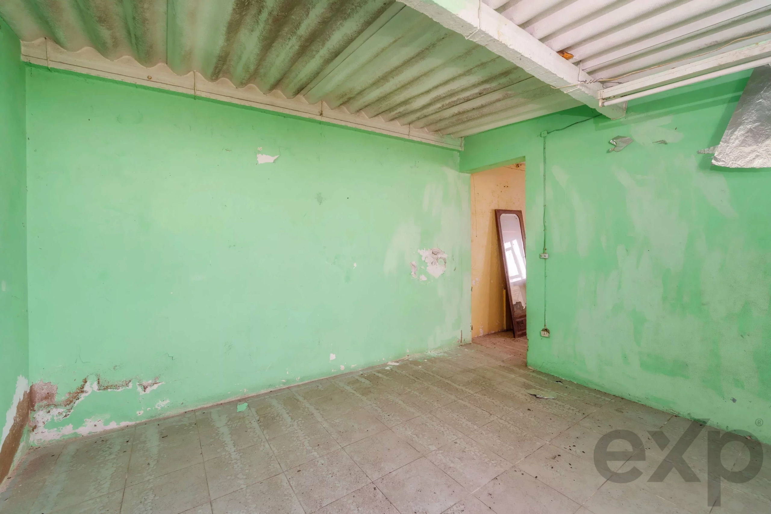 Empty green-walled room with peeling paint and a doorway revealing another space; a mirror leans against the doorway.