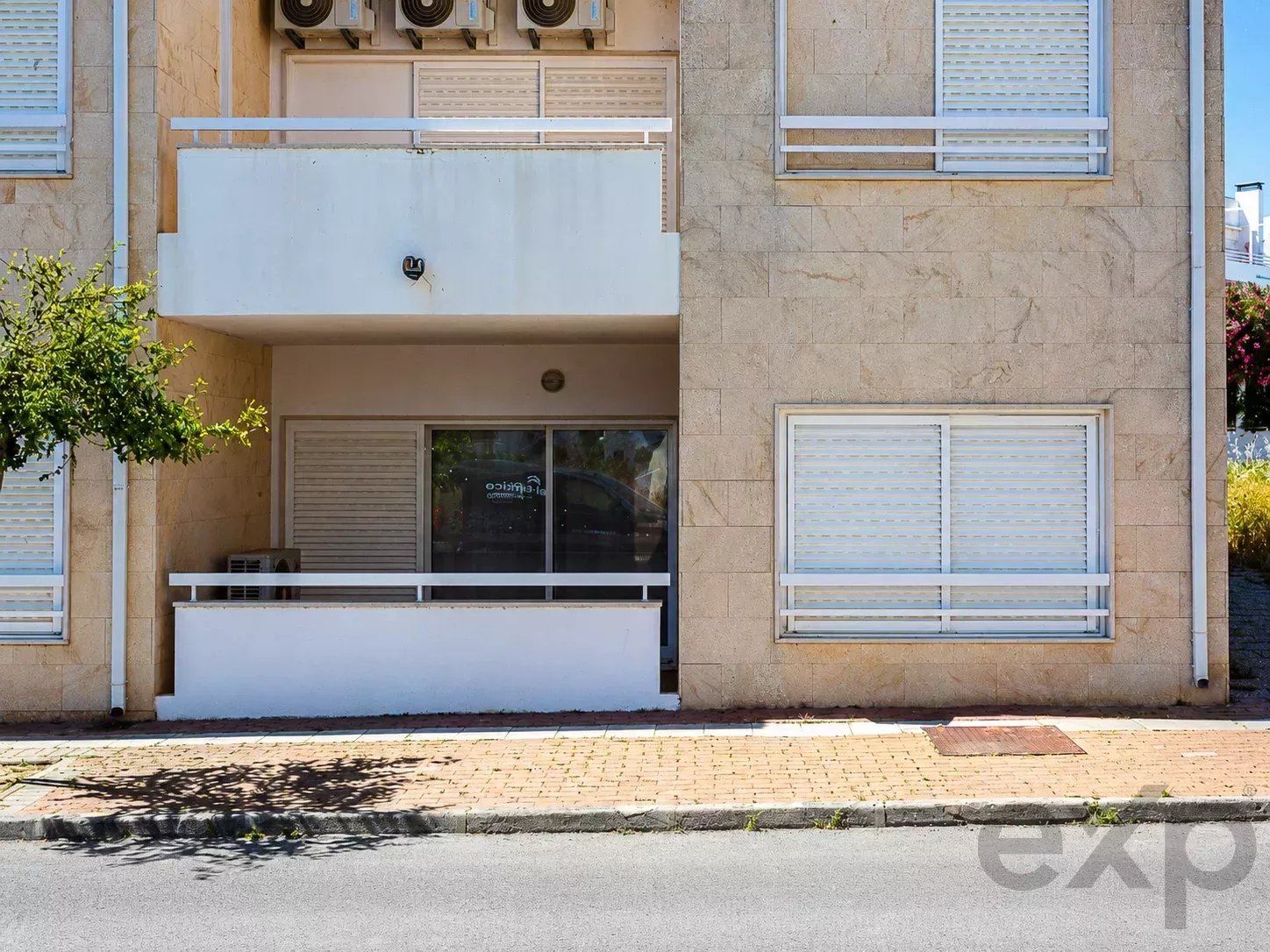 Ground-floor storefront with a glass door and window blinds, beige stone building, and a small front balcony above.