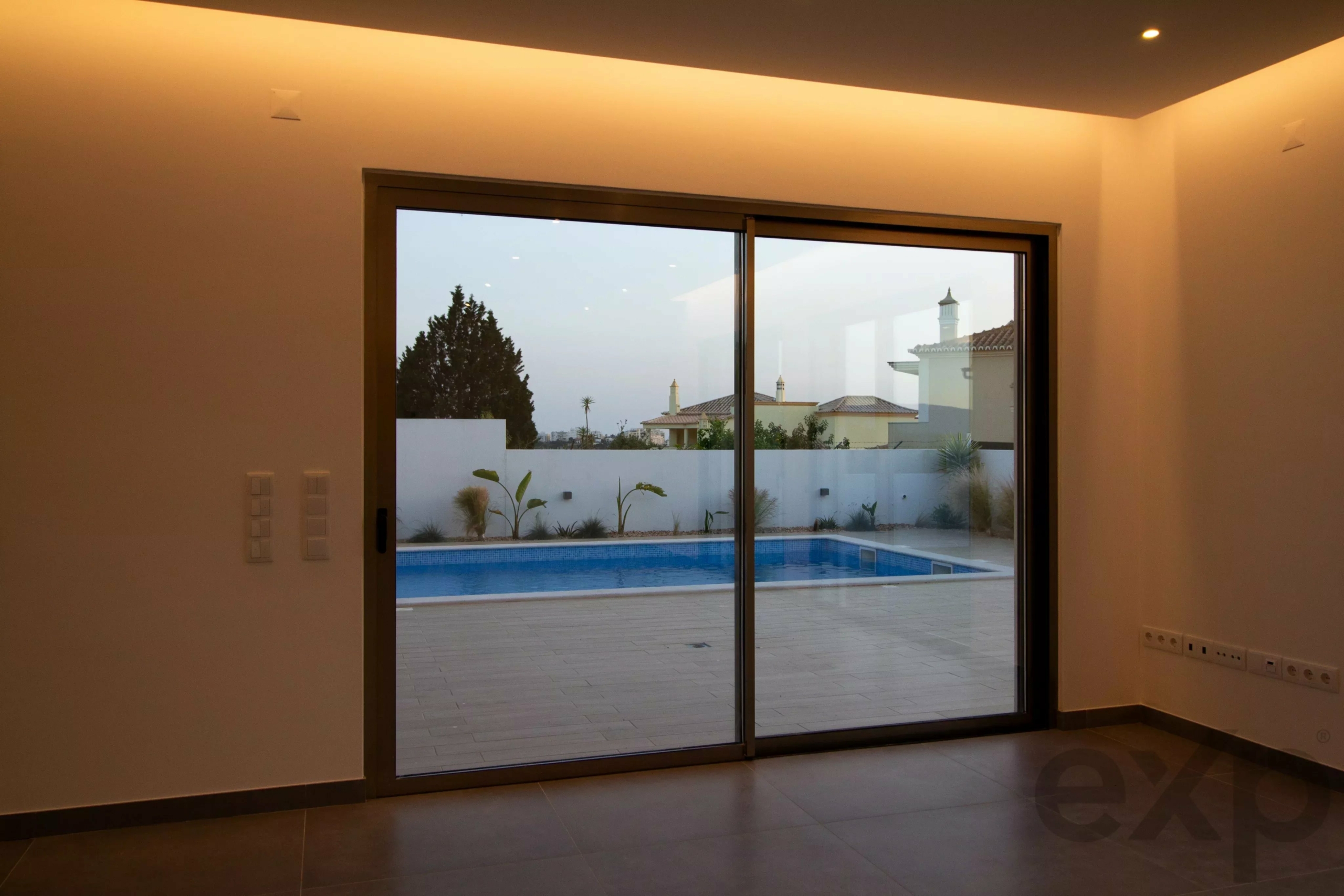 Interior room with large sliding glass doors opening to a pool, warm overhead lighting, and a fenced yard visible outside.