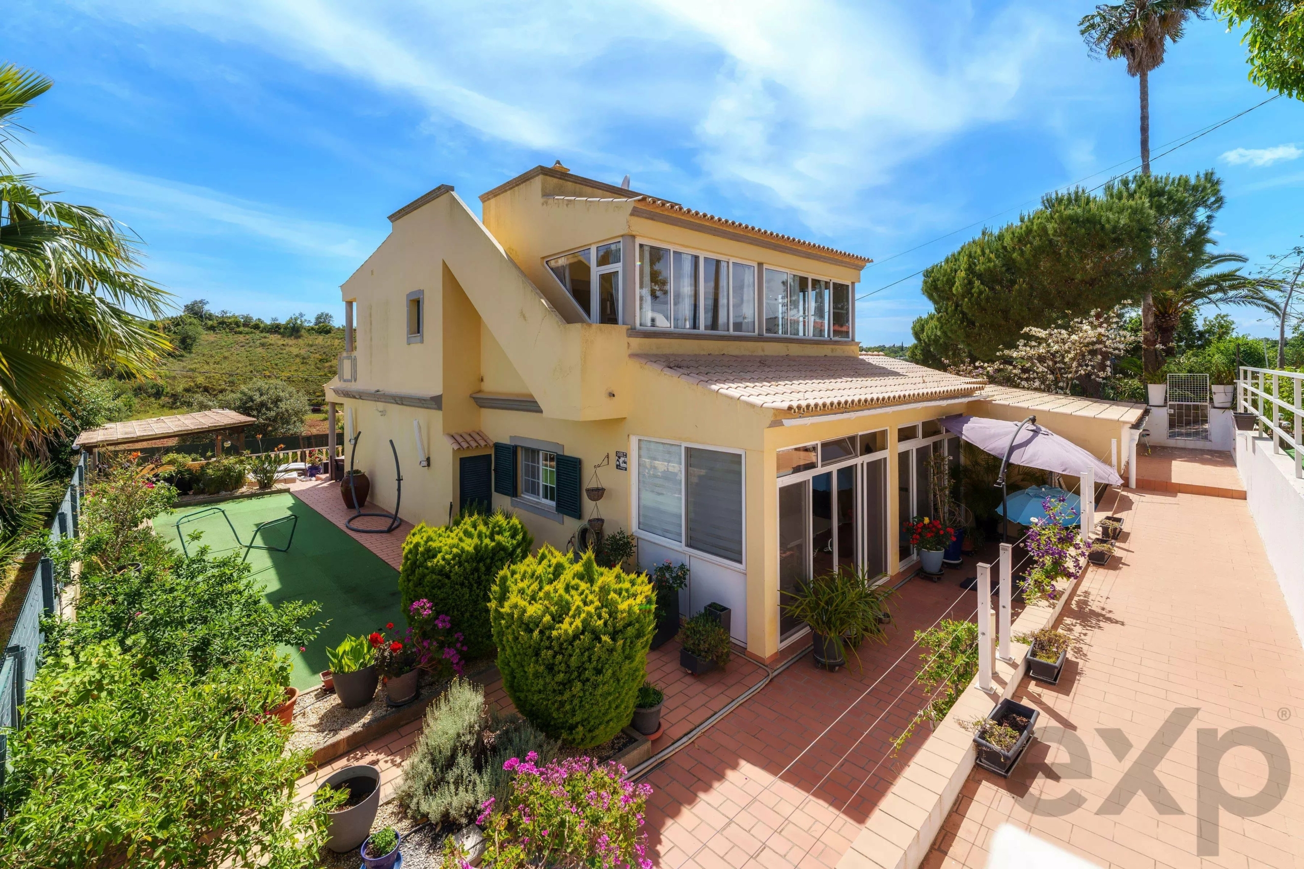 Two-story yellow house with a tiled roof, glass-enclosed balcony, and a lush garden on a sunny day