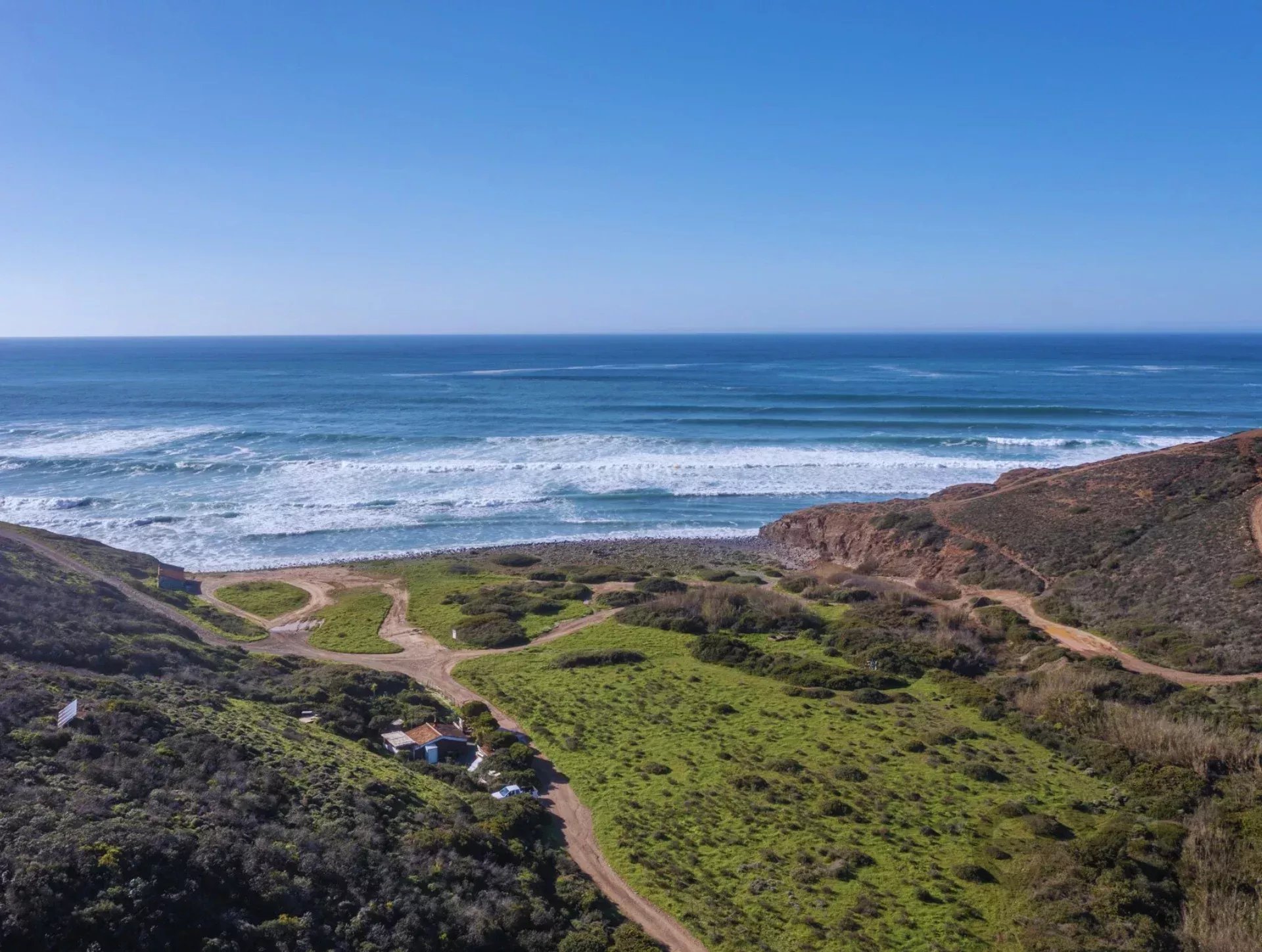 A coastal hillside with winding dirt roads, small houses, and green grassy slopes leading to the ocean horizon under a clear blue sky