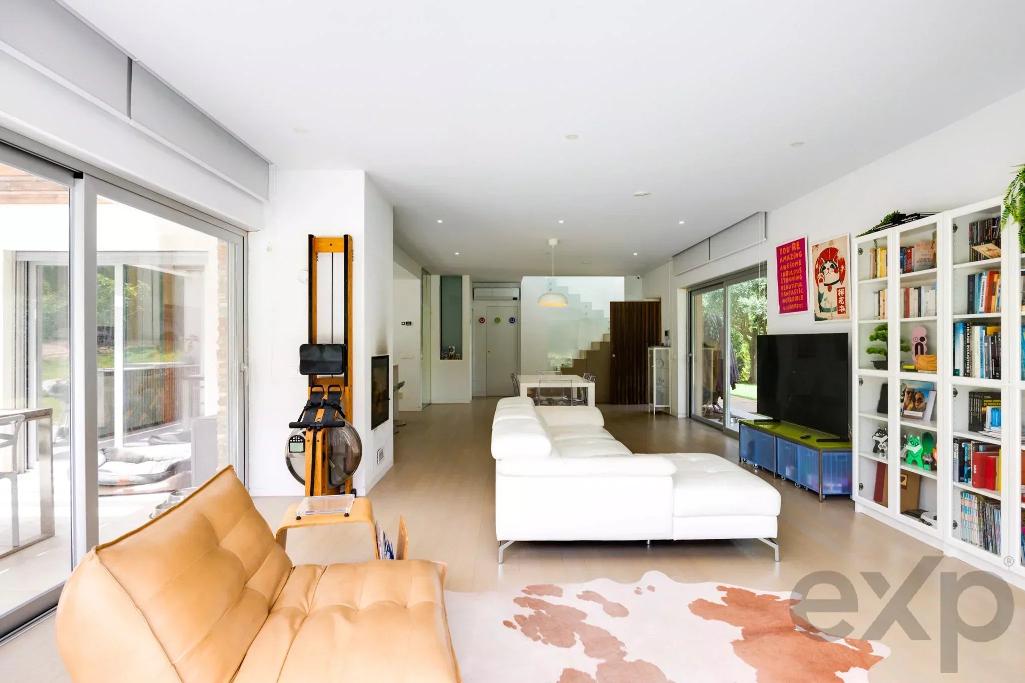 Bright open-plan living room with white sofas, wooden chairs, and a large glass wall system opening to outdoors; bookshelves line the right wall.