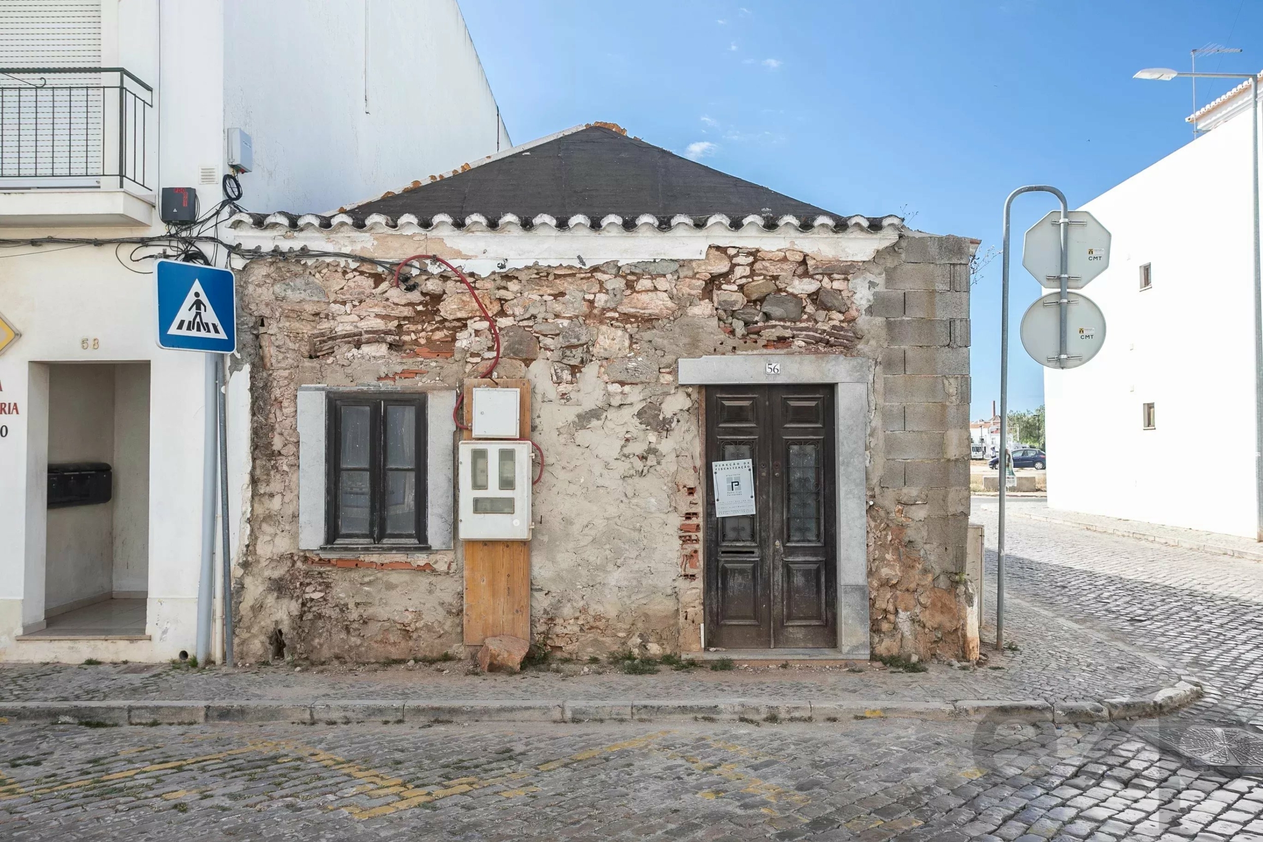 Old stone building with worn, partially demolished facade and dark wooden door on a cobblestone street under a blue sky