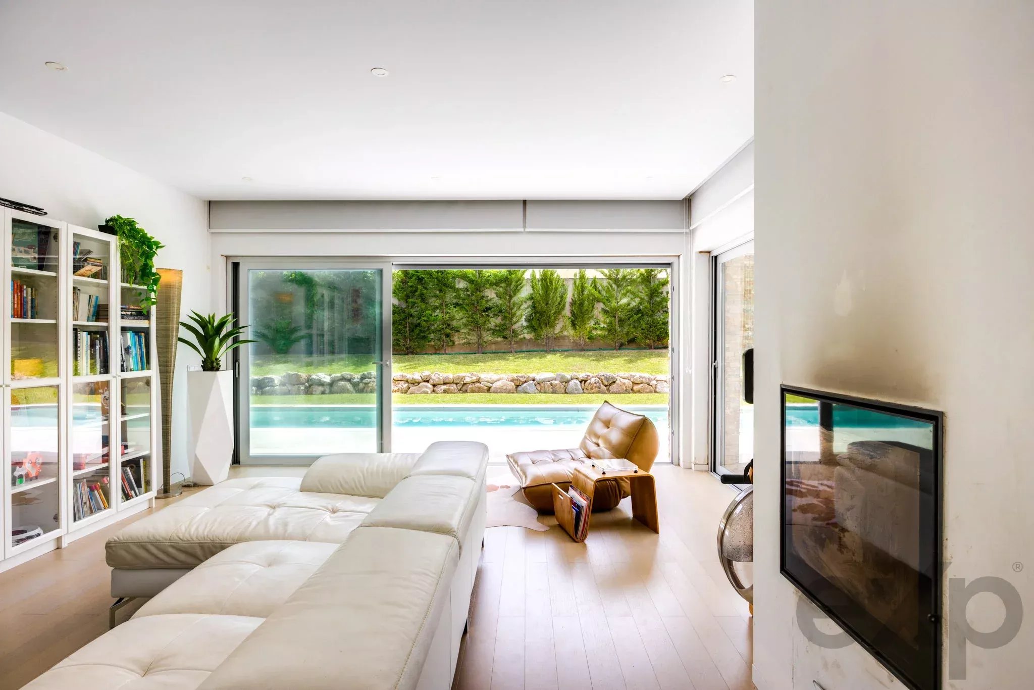 Bright modern living room with a white sectional sofa, glass-front bookcases, and potted plants, opened to a pool and garden area outside.