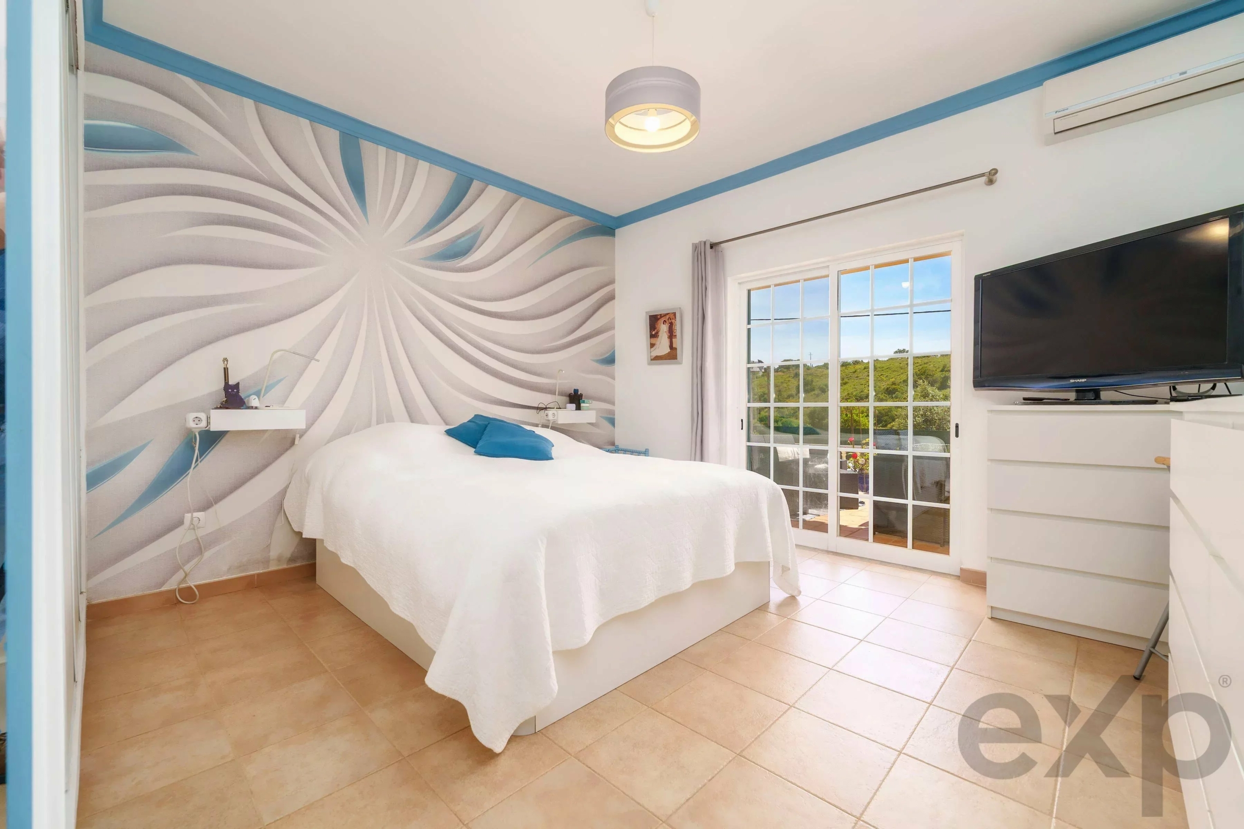 Bright bedroom with abstract blue and white swirl mural, white bed with blue pillows, and a large window showing greenery outside.