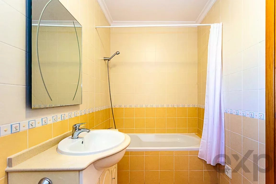 Bathroom interior with a white sink, oval mirror, and yellow-tiled walls surrounding a tub and handheld shower head. A white shower curtain hangs at the tub.