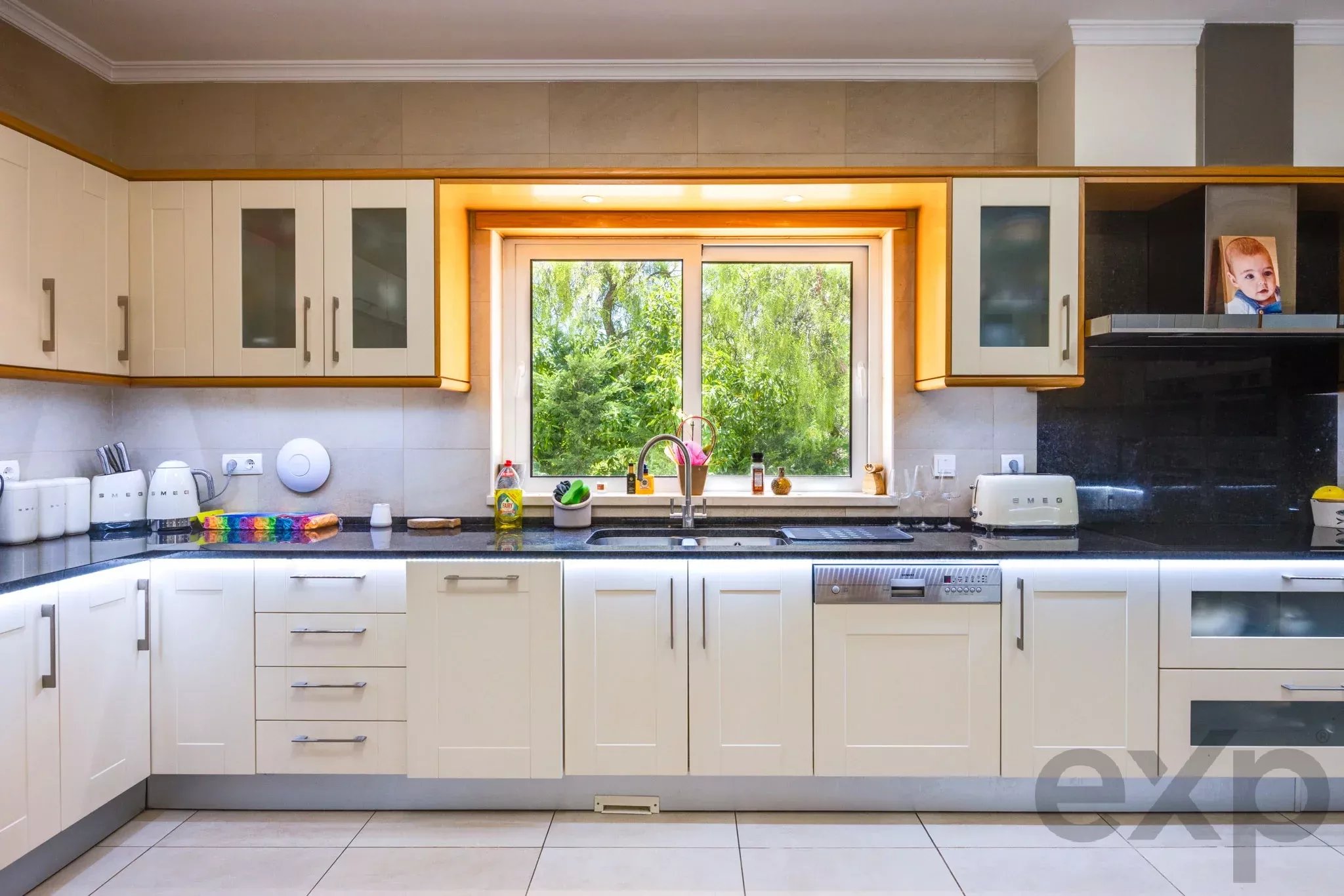 Bright modern kitchen with white cabinets, black countertops, and a central window above the sink overlooking greenery.