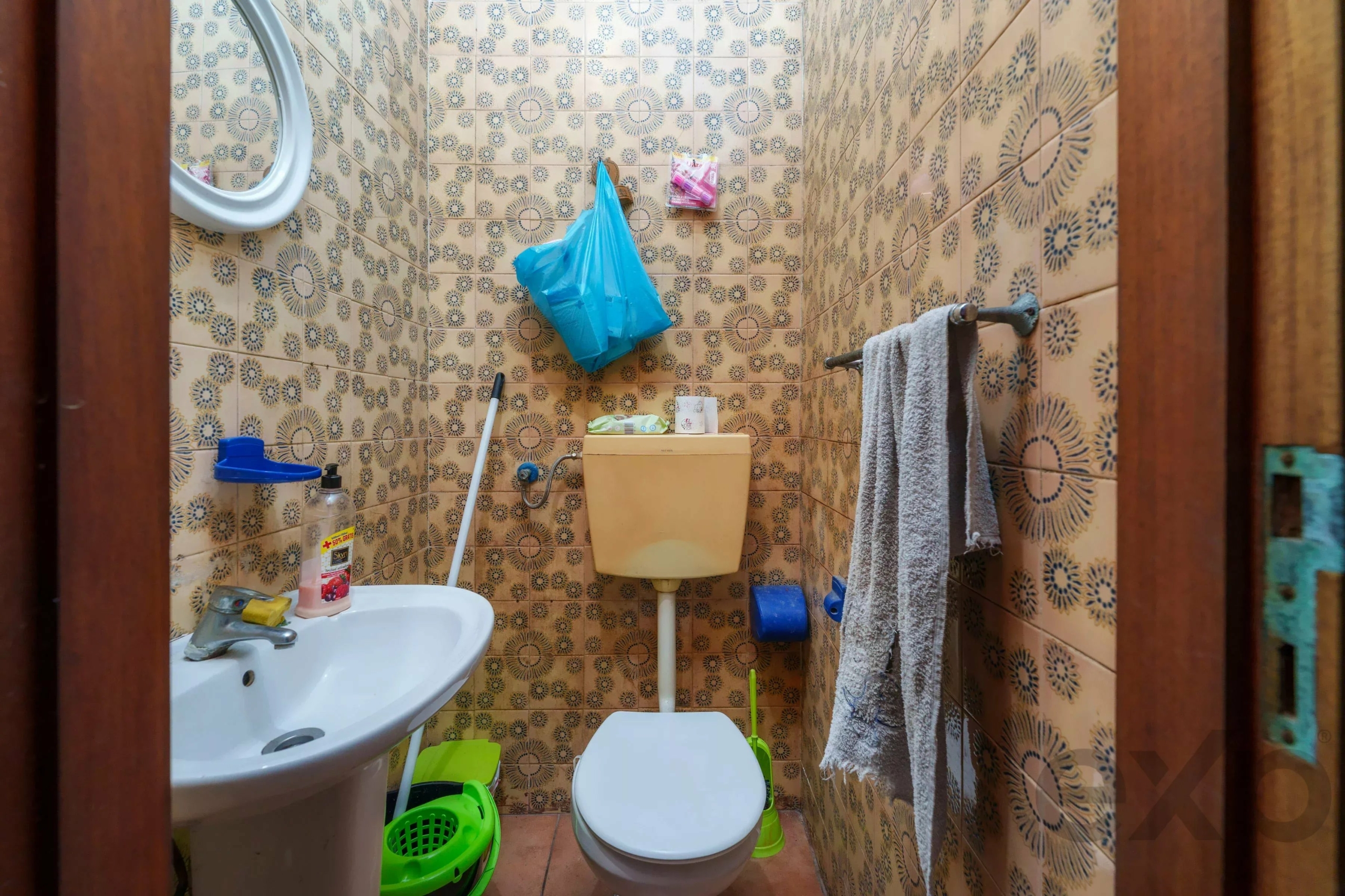 Small bathroom with patterned tiled walls, a beige toilet, white sink, gray towel on a rack, and a blue plastic bag hanging on the wall above the toilet.